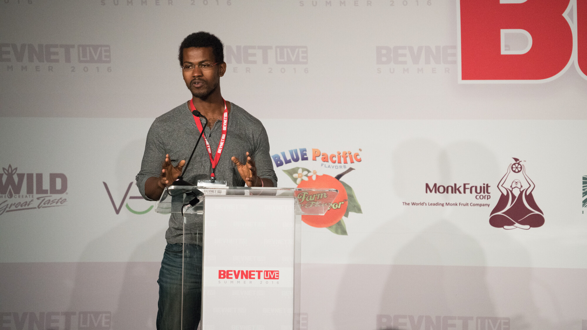 Man in glasses and a grey t-shirt with a BevNET Live lanyard at a clear podium, with Wild, Blue Pacific and Monk Fruit Corp logos on the backdrop.
