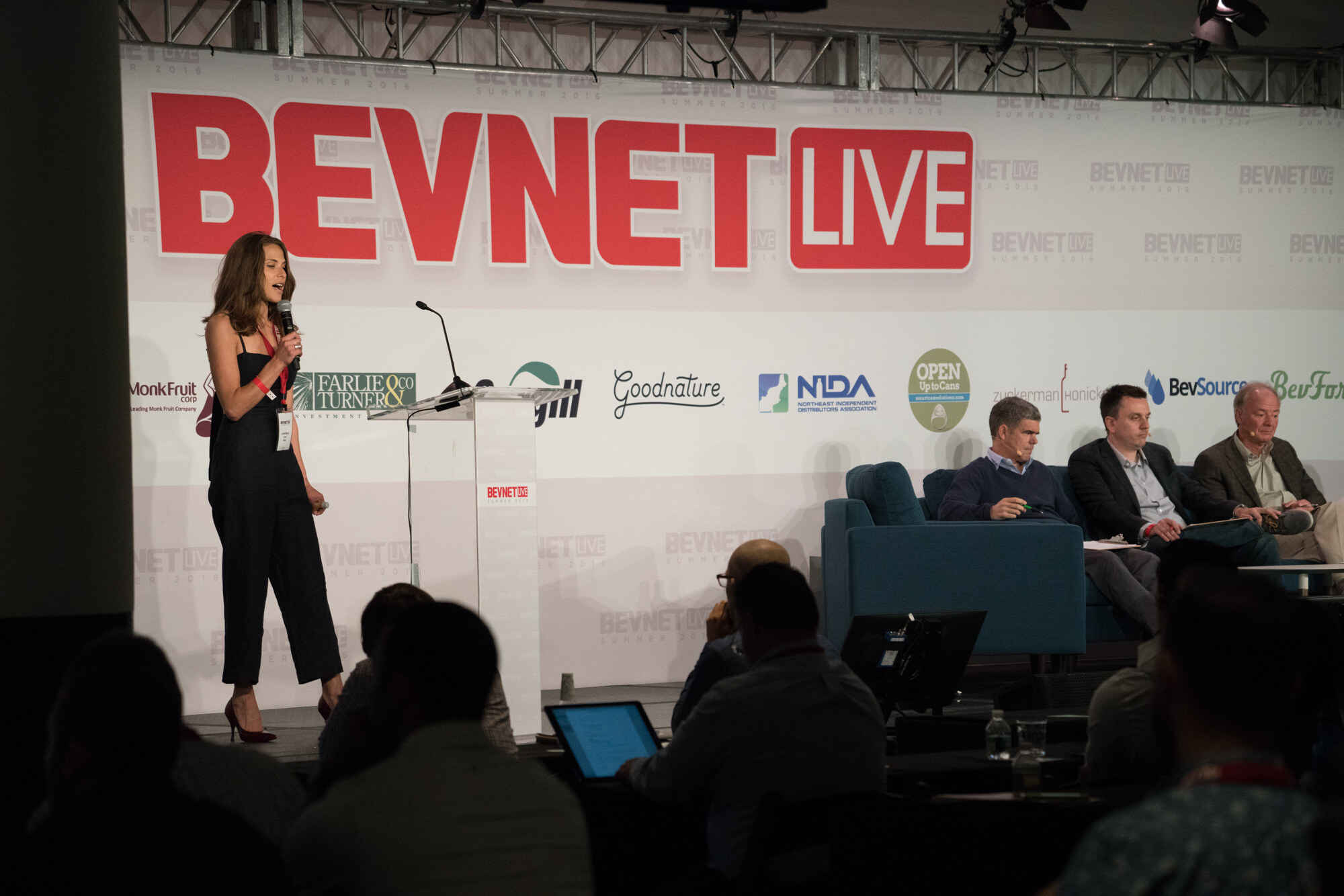 Woman in a black jumpsuit and BevNET Live lanyard holding a microphone beside a clear podium, with three men on a sofa at right and audience heads and a laptop in the foreground.