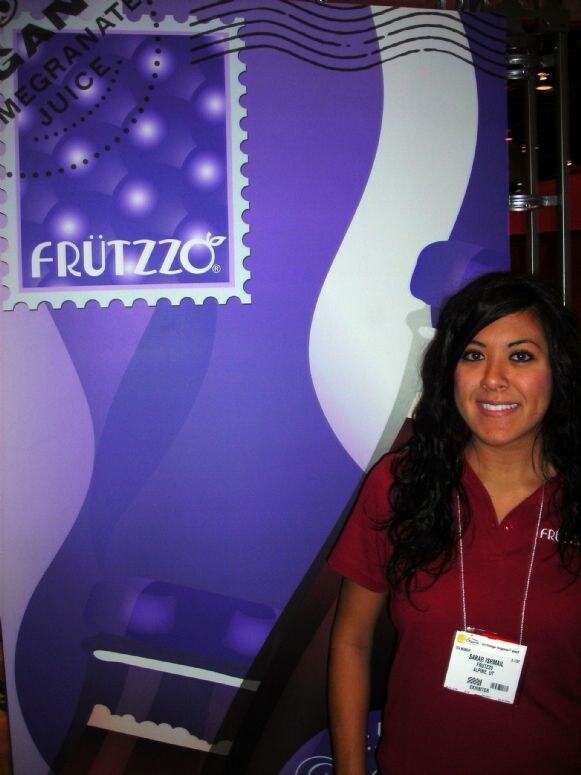 Sarah Ishmail of Frutzzo, with long dark hair and a maroon shirt, smiles beside a purple Frutzzo pomegranate juice banner designed like a postage stamp.