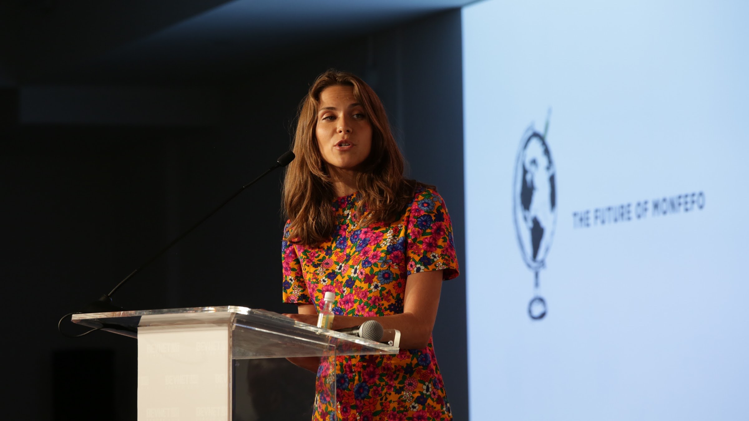 Woman with long brown hair in a colorful floral dress at a clear acrylic podium with a microphone, a projection reading The Future of Monfefo beside her.