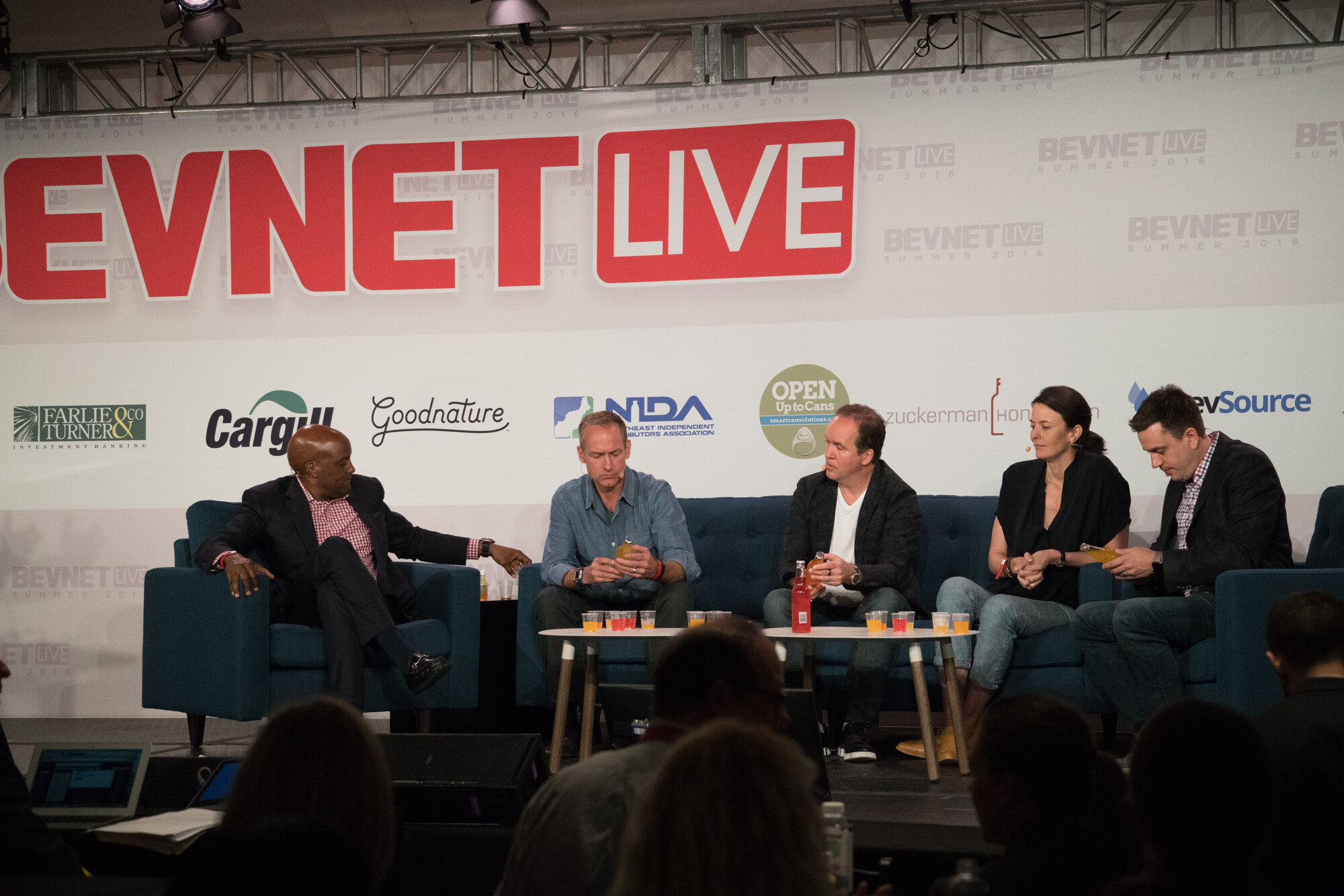 Wide stage view of five people on blue seats around two low white tables with small orange and red tasting cups, under the BevNET Live sign, with audience heads in the foreground.