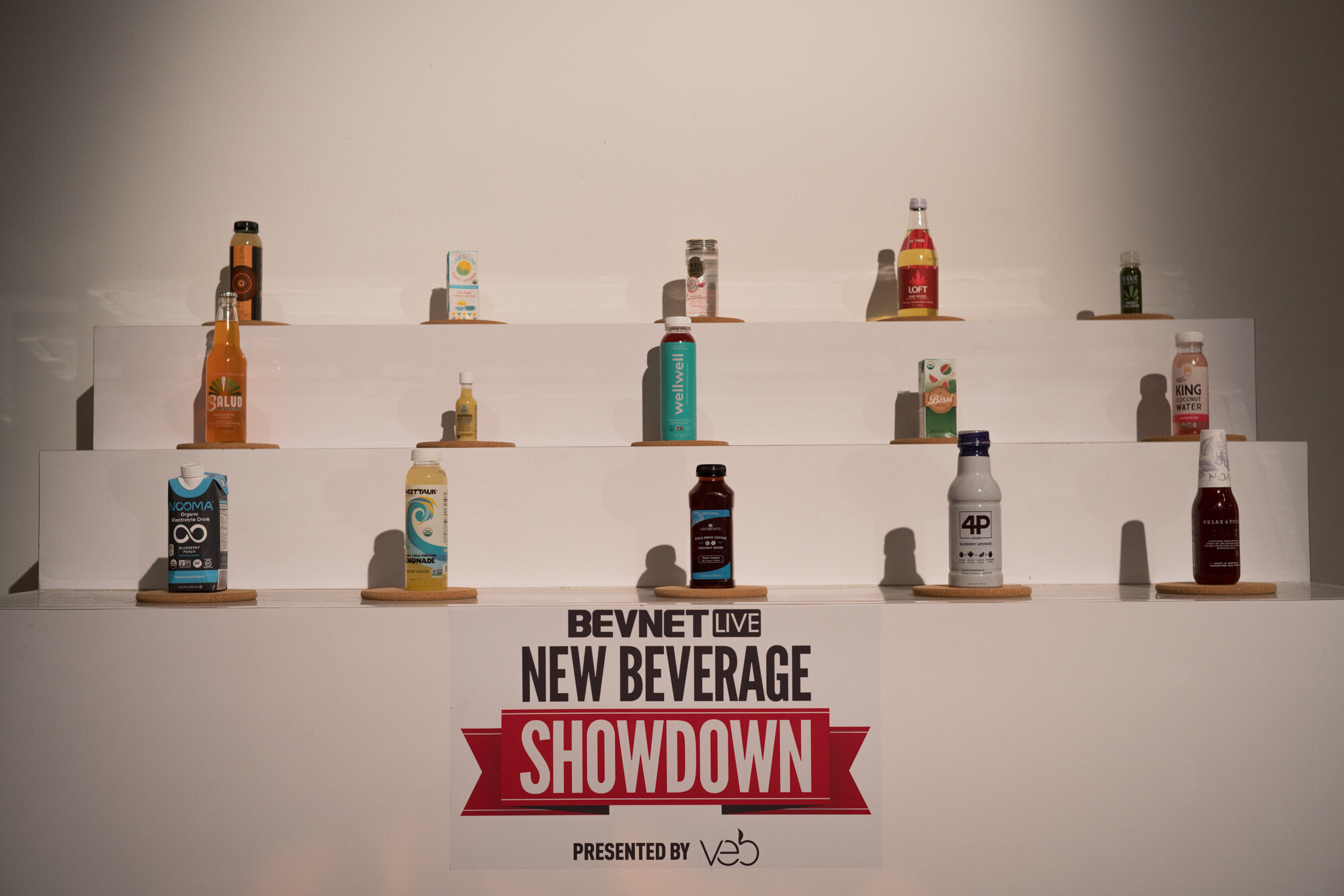 Three tiers of white shelving with beverage bottles and cartons on cork coasters, including wellwell, Salud, King Coconut Water, 4P and LOFT, above a sign reading BevNET Live New Beverage Showdown presented by Veb.