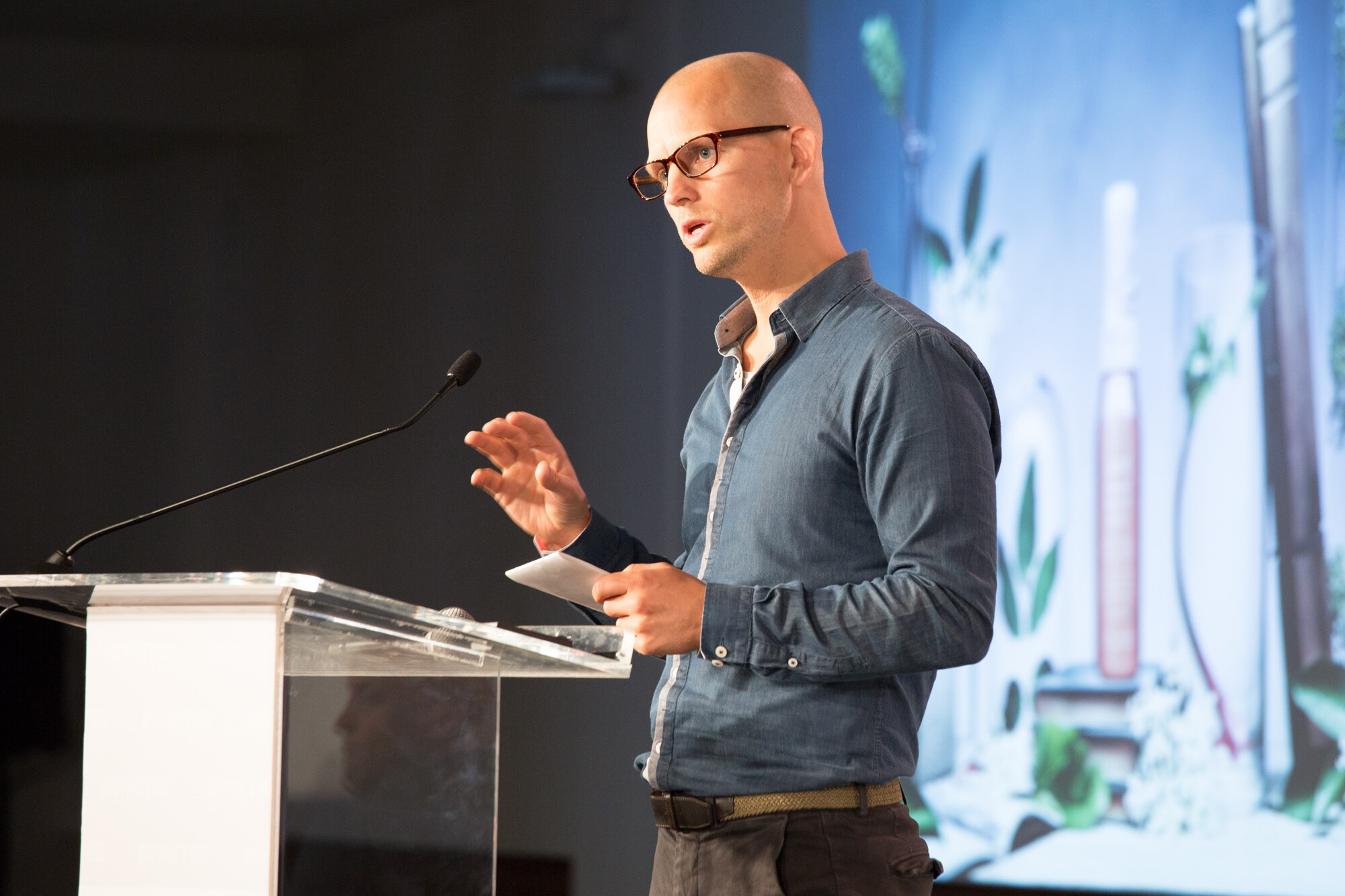 Bald man in dark-framed glasses and a blue chambray shirt at a clear podium holding a card, with a projected image of bottles and green leaves behind him.