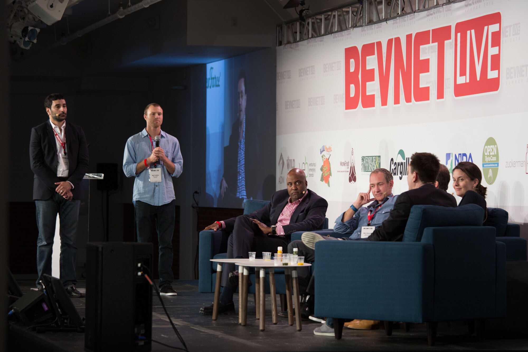 Wide stage view of two standing men, one holding a microphone, beside judges on blue armchairs and sofas under a red BevNET Live sign, with a video screen showing a close-up at left.