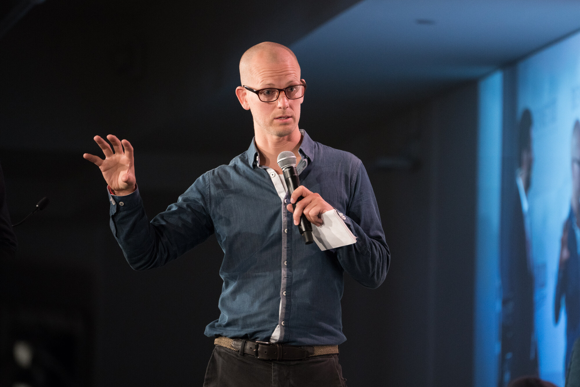 Man in dark-framed glasses and a blue chambray shirt holding a microphone, with a blue-lit screen at the edge of the frame.