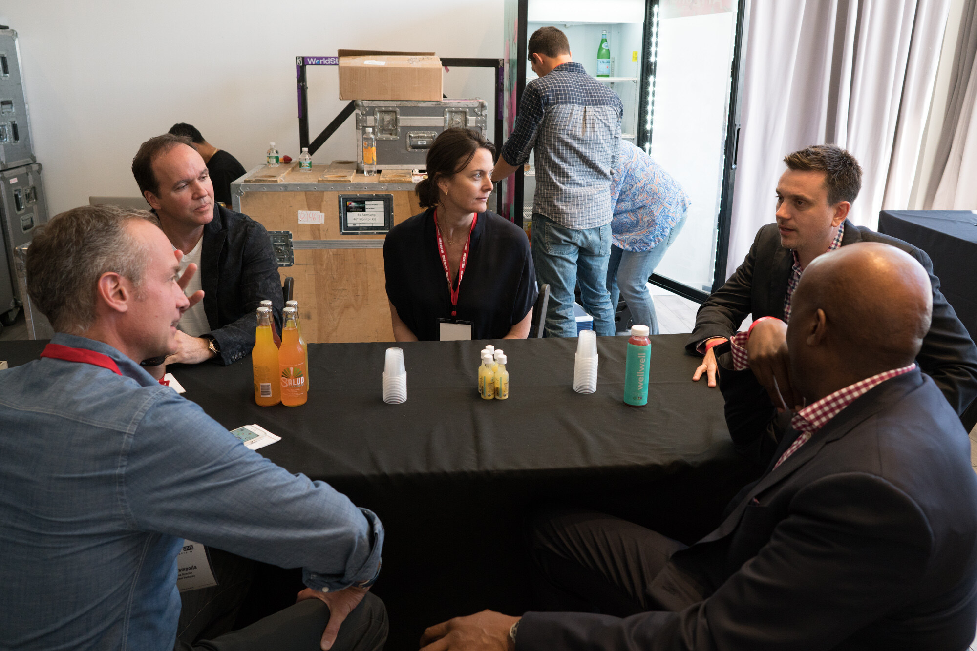 Five people around a black-draped table with orange Salud bottles, small shot bottles and a teal wellwell bottle, with road cases and a stagehand in a plaid shirt behind them.