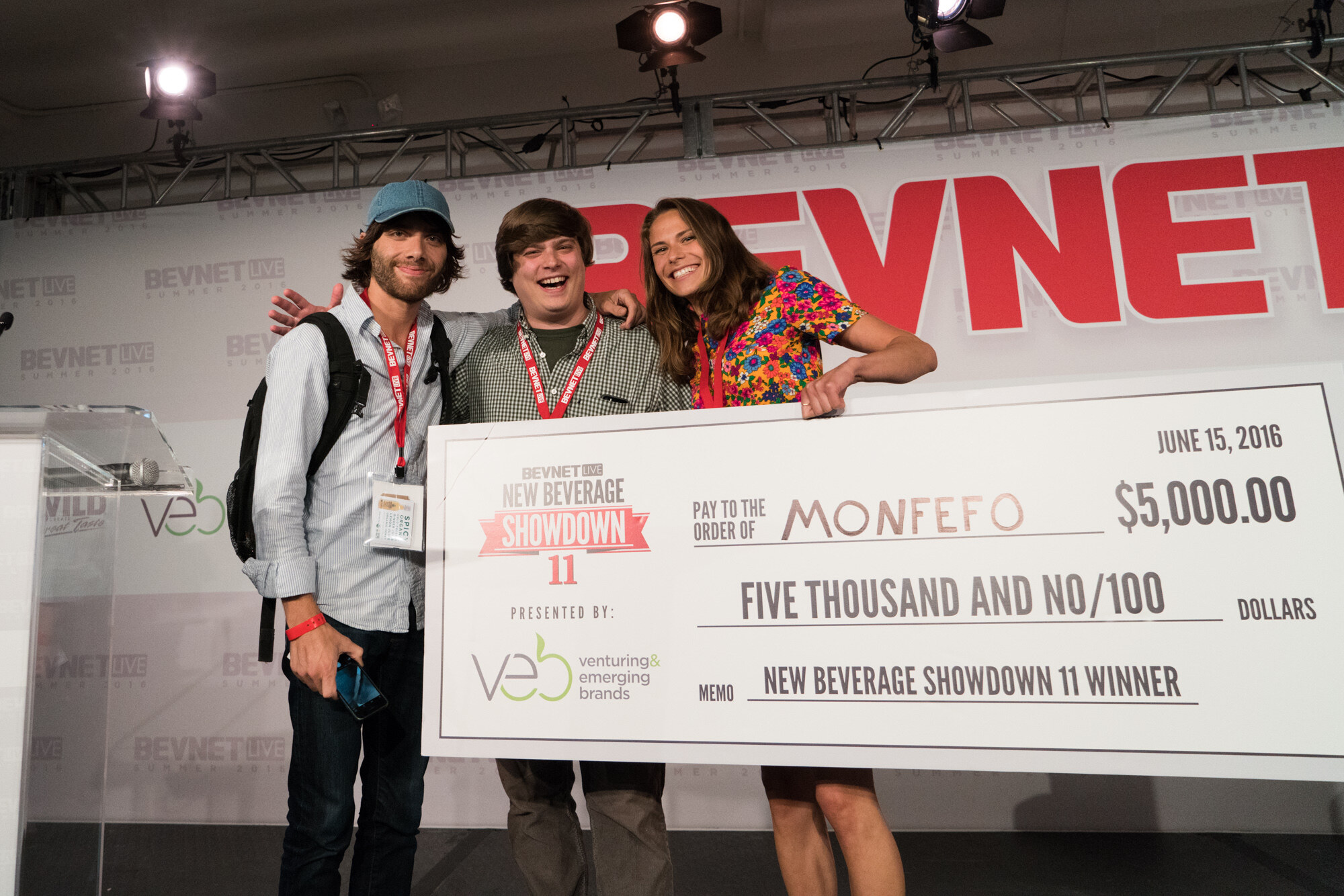 Two men and a woman in a floral dress laughing behind a large check dated June 15, 2016, reading Pay to the order of Monfefo, $5,000.00, New Beverage Showdown 11 winner, presented by Venturing & Emerging Brands.