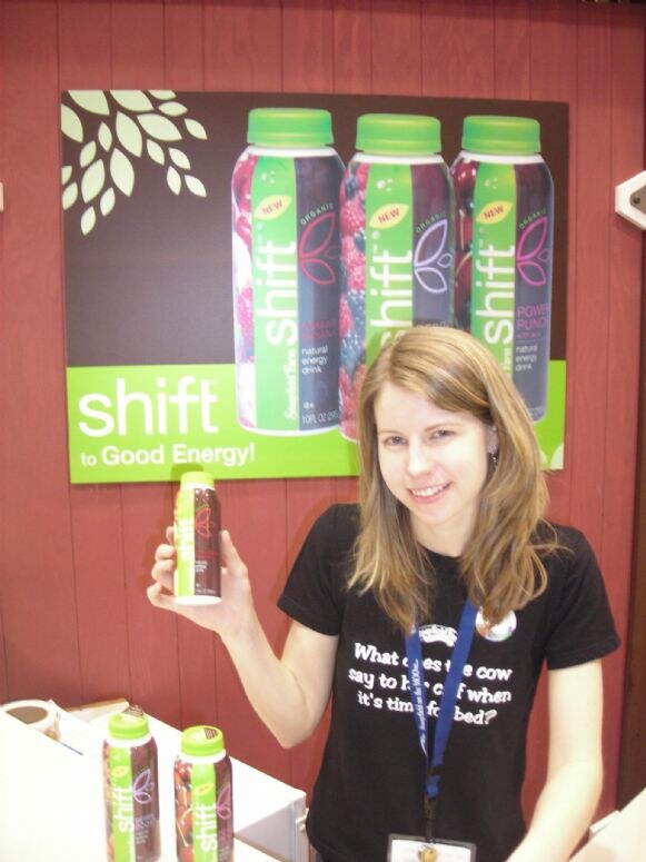 A smiling woman in a black T-shirt holds up a bottle of shift energy drink in front of a poster reading shift to Good Energy. More shift bottles sit on the counter in front of her.