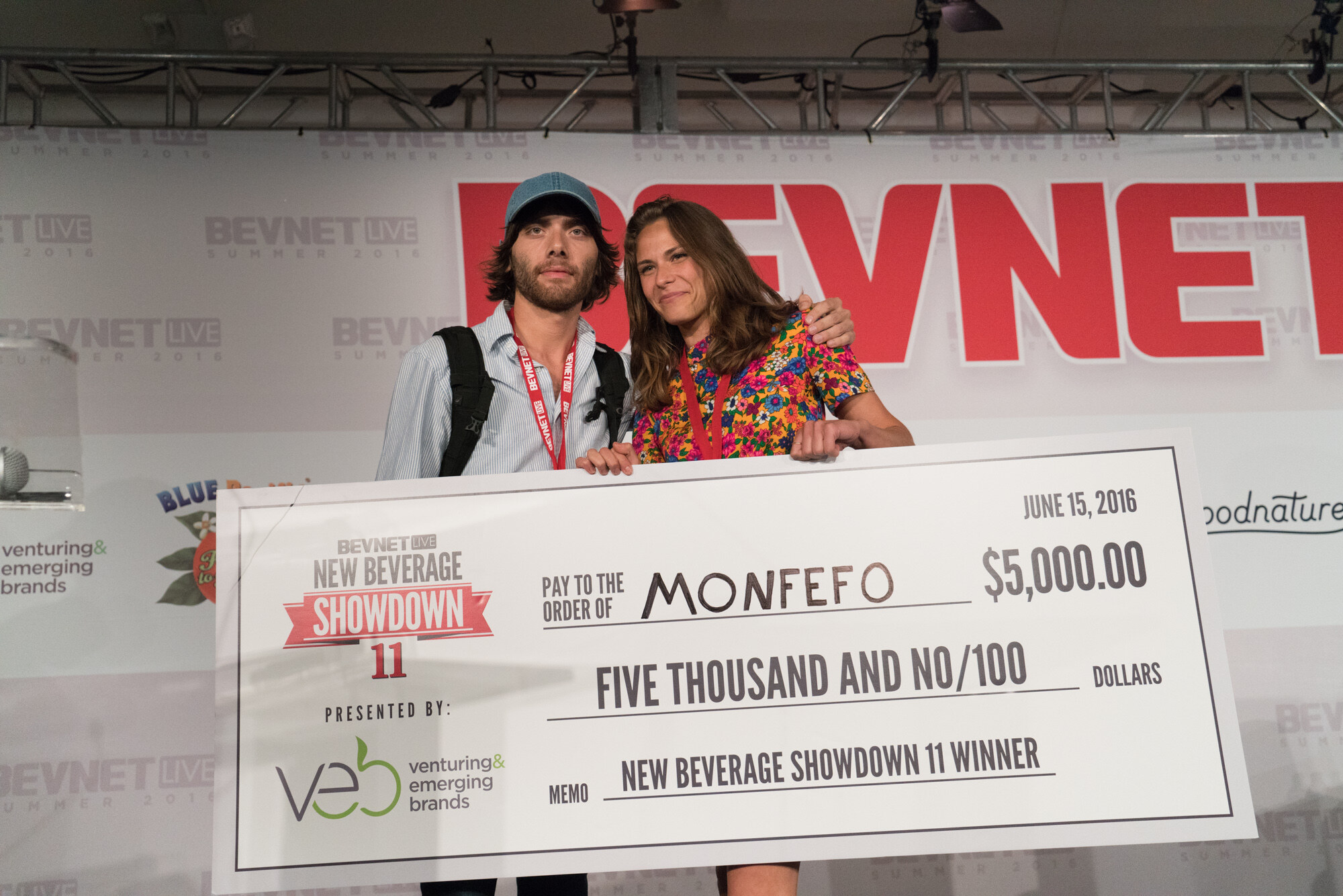 Man in a blue cap and backpack and woman in a floral dress behind a large check reading Monfefo, $5,000.00, New Beverage Showdown 11 winner, in front of the BevNET Live backdrop.
