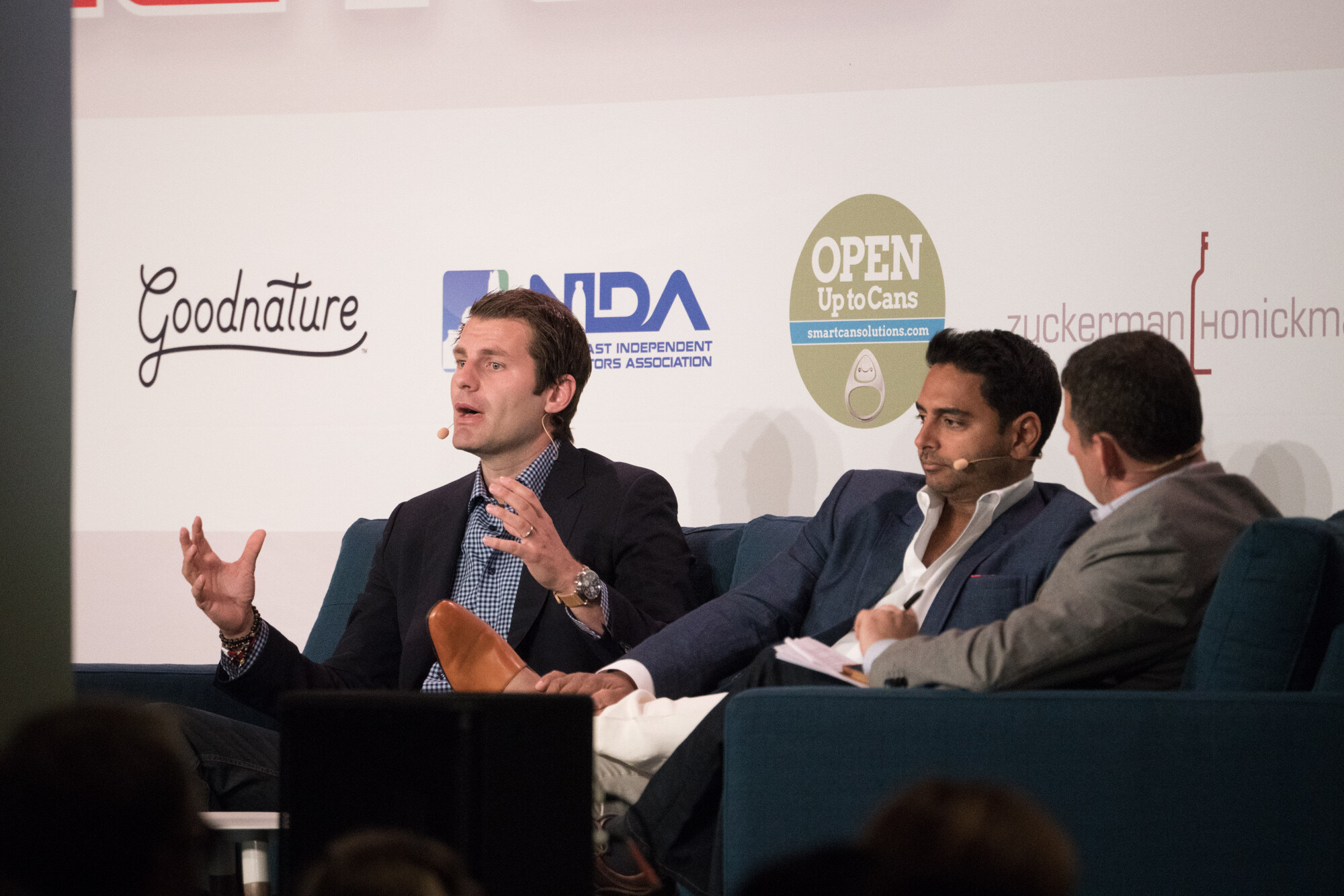 Three men on a blue sofa on stage, the one at left in a dark blazer and checked shirt gesturing, in front of Goodnature, NIDA and Open Up to Cans logos.