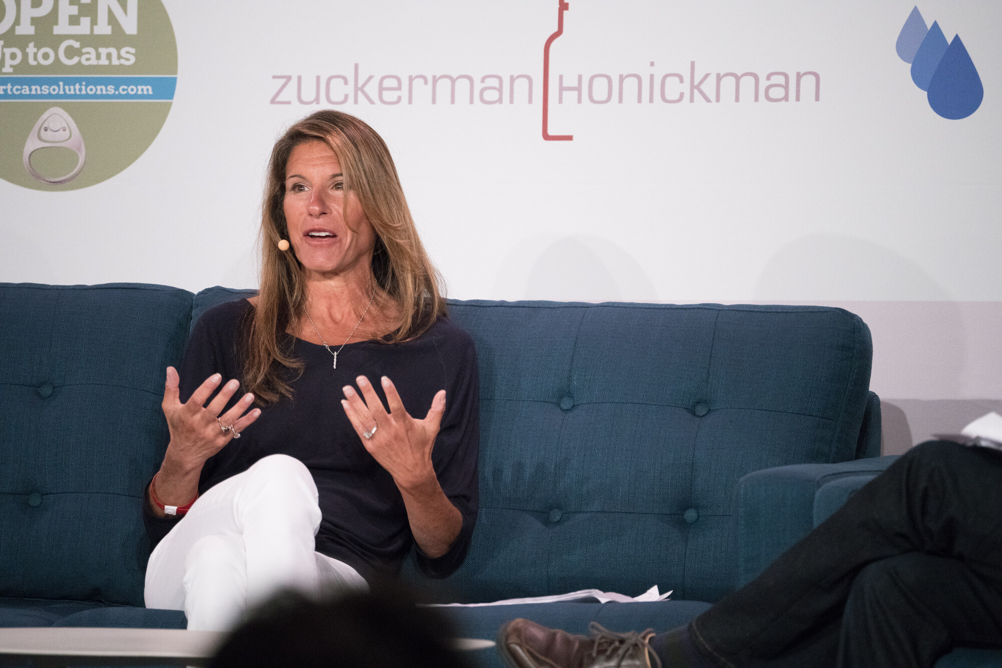 Woman with long brown hair and a headset mic seated on a blue sofa with her hands open, in front of Open Up to Cans and Zuckerman Honickman logos.