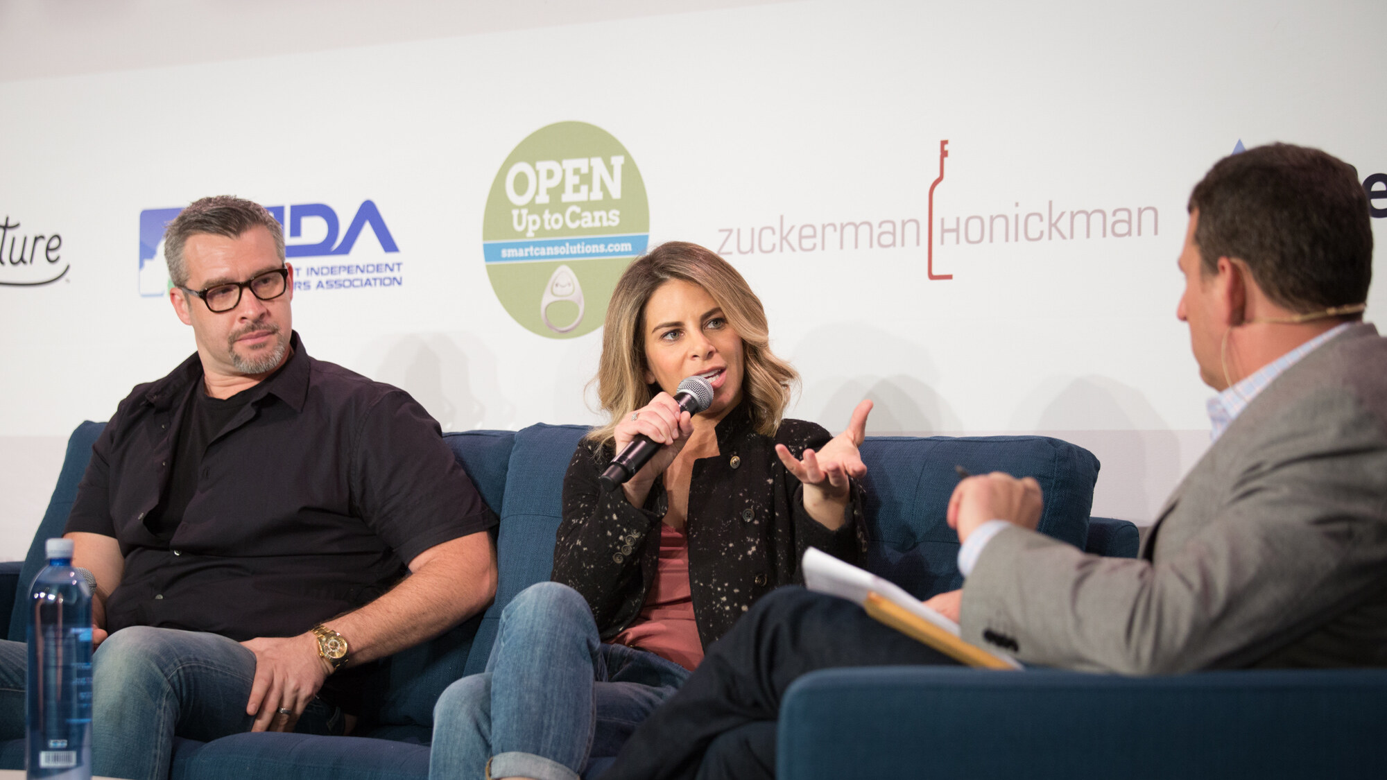 Three people on a blue stage sofa: a man in glasses and a black shirt, a woman in a dark jacket speaking into a microphone, and an interviewer in a grey blazer holding notes, with Open Up to Cans and Zuckerman Honickman logos behind.