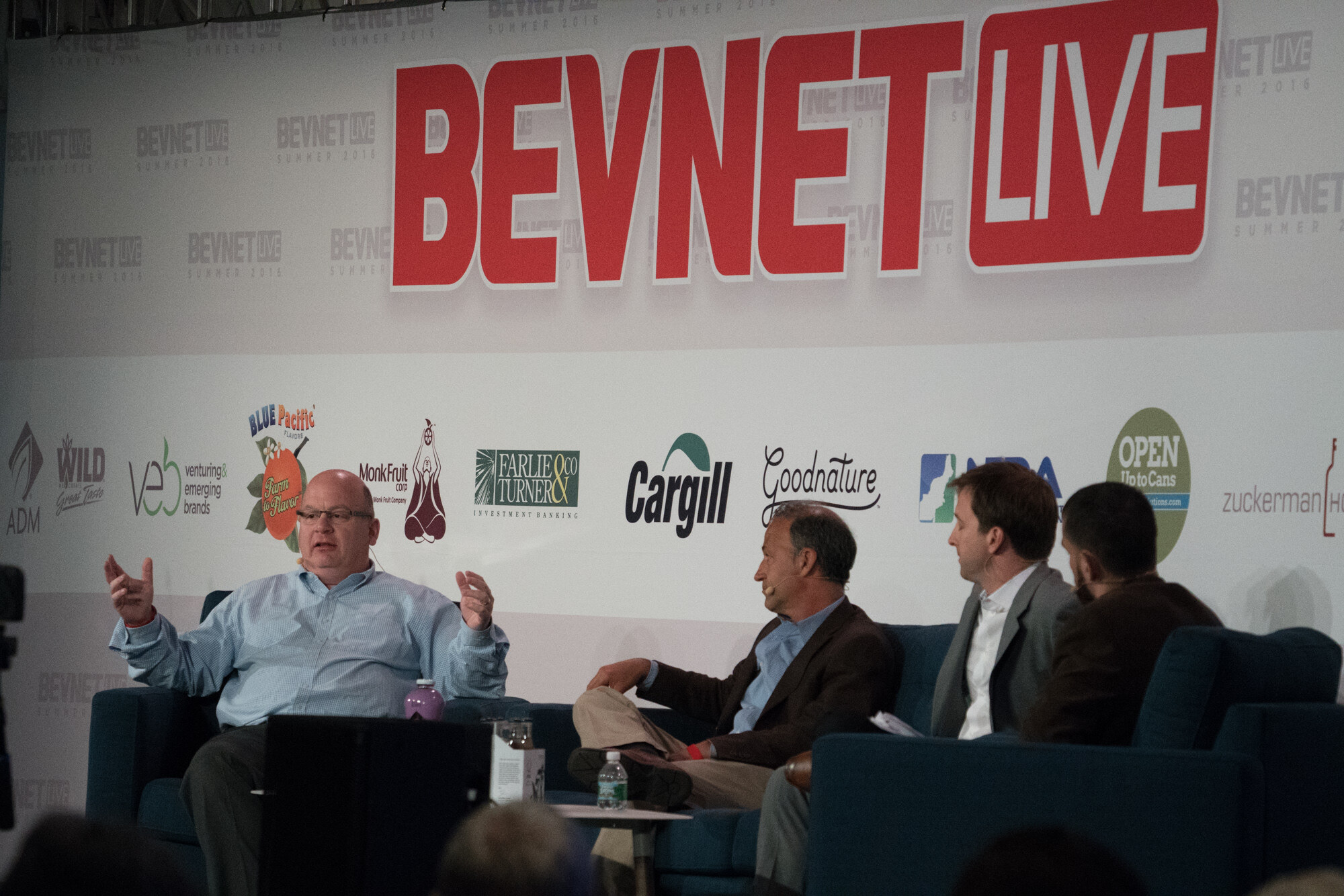 Four men on stage beneath the red BevNET Live sign and a row of sponsor logos, the man at left in a light blue shirt gesturing as he speaks.