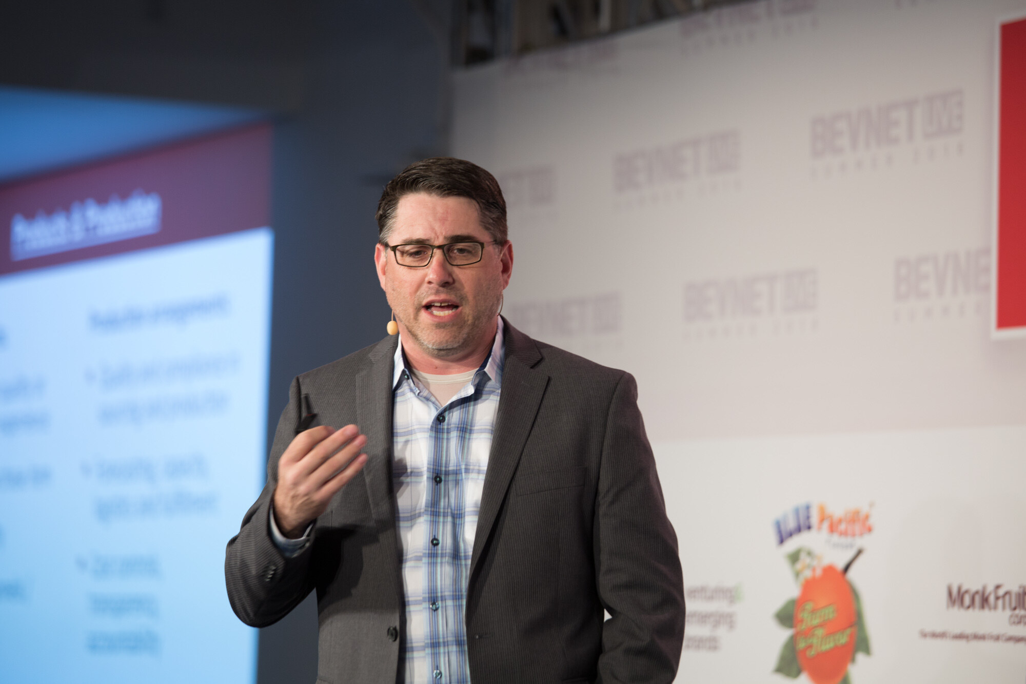 Man in glasses, a grey blazer and a blue plaid shirt with a headset mic, speaking next to a blurred projected slide, with Blue Pacific and Monk Fruit Corp logos behind him.