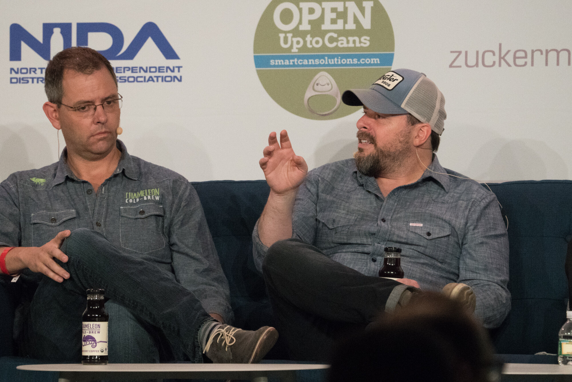 Two men on a blue sofa, one in glasses wearing a Chameleon Cold-Brew shirt, the other in a trucker hat gesturing, with Open Up to Cans and NIDA signs behind them.