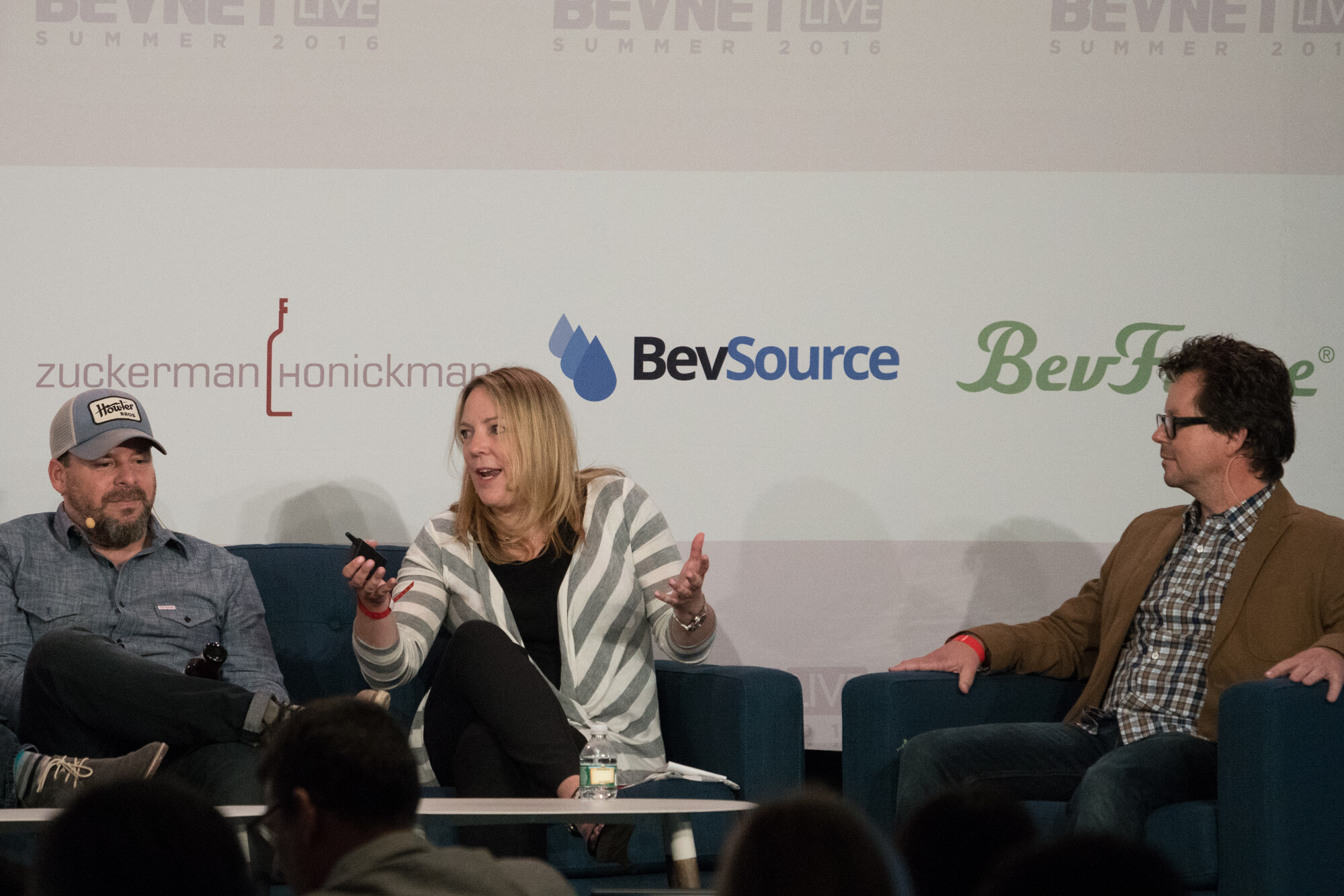 Woman in a grey striped cardigan holding a clicker, a man in a trucker hat at left and a man in a brown blazer and glasses at right, in front of BevSource and BevForce logos.