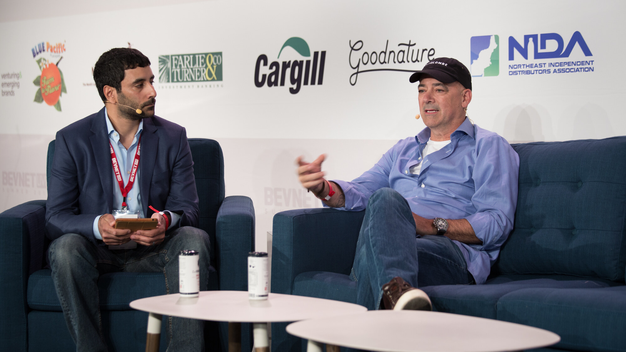 Two men on stage seats, one in a navy blazer with a red lanyard and one in a black cap and blue shirt, with two small cans on white tables and Cargill, Goodnature and NIDA logos behind.