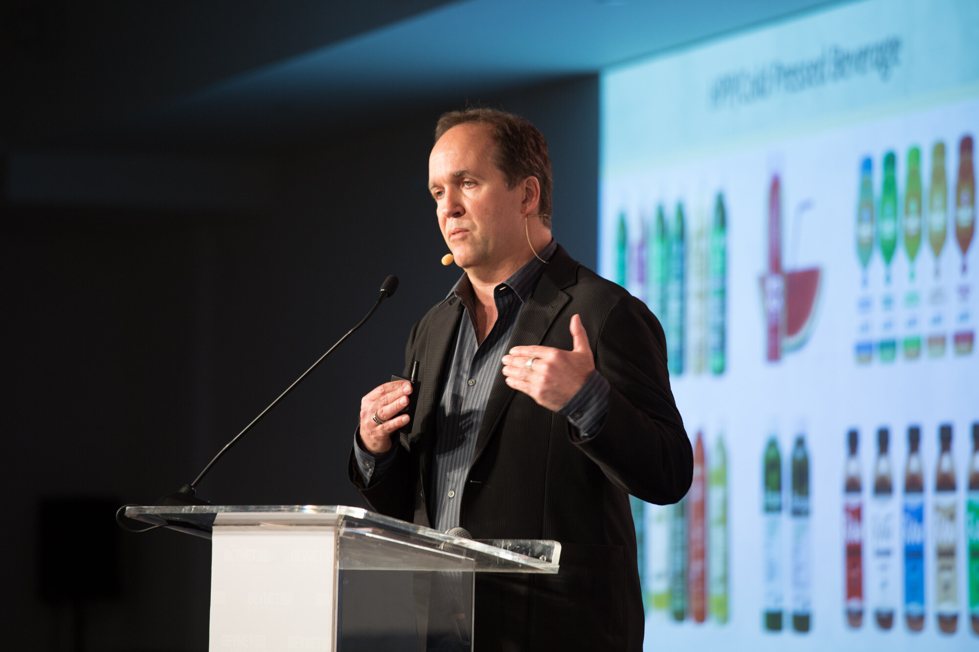 Man in a black blazer and striped shirt with a headset mic at a clear acrylic podium, a projected slide titled HPP/Cold Pressed Beverage showing rows of bottles behind him.