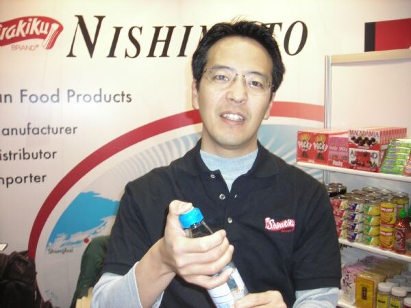 A man with glasses in a black Shirakiku polo holds a water bottle in front of a Nishimoto banner and shelves of Pocky and other Japanese food products.