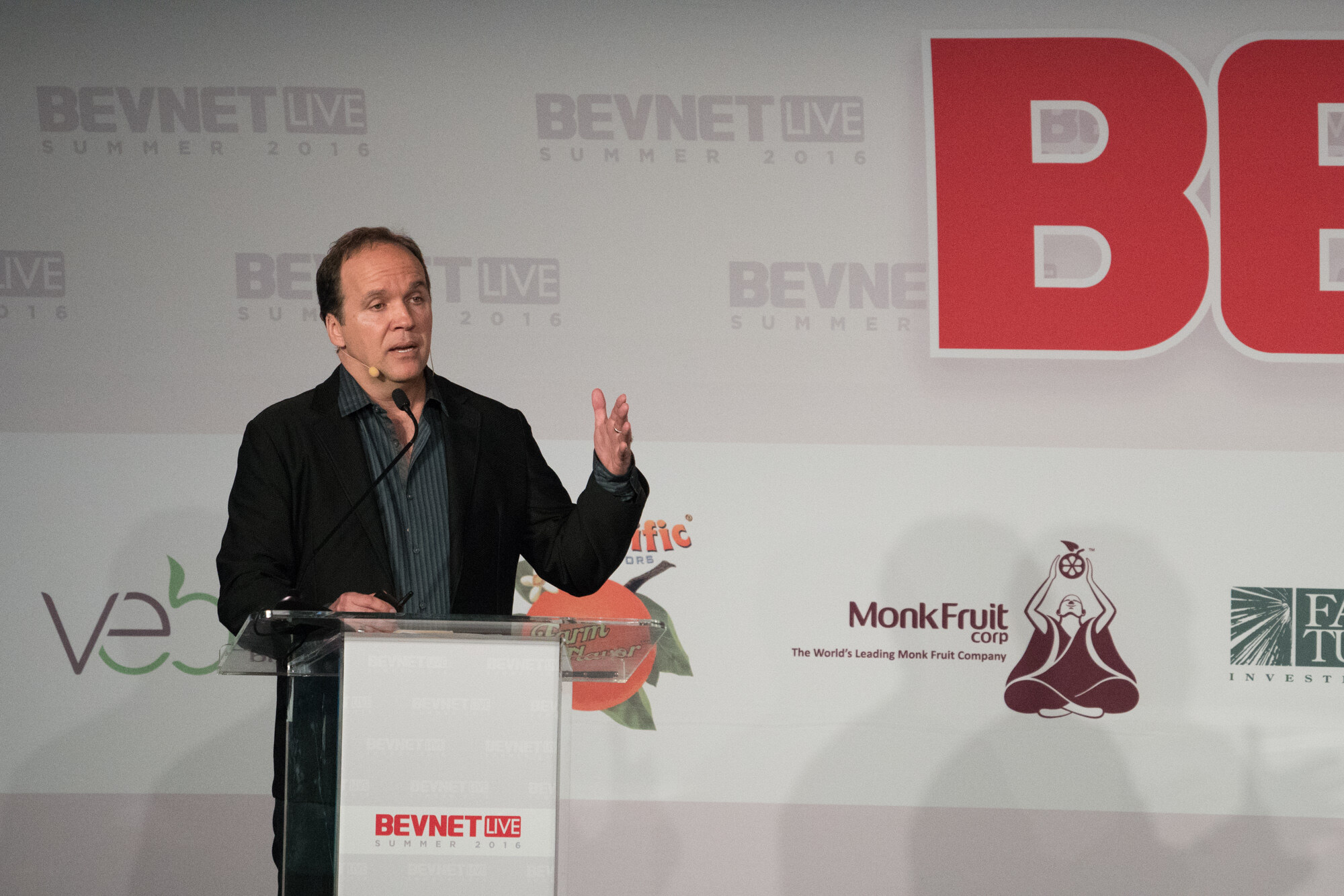 Man in a black blazer over a striped shirt with a headset mic at a podium reading BevNET Live Summer 2016, with Monk Fruit Corp and Veb logos on the backdrop.