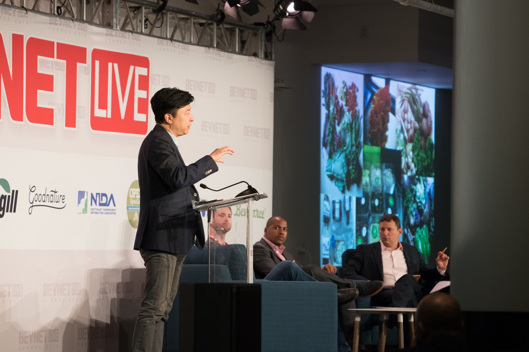 Man in a navy blazer standing at a clear podium, a large screen filled with photos of vegetables behind him and three seated panelists at right.