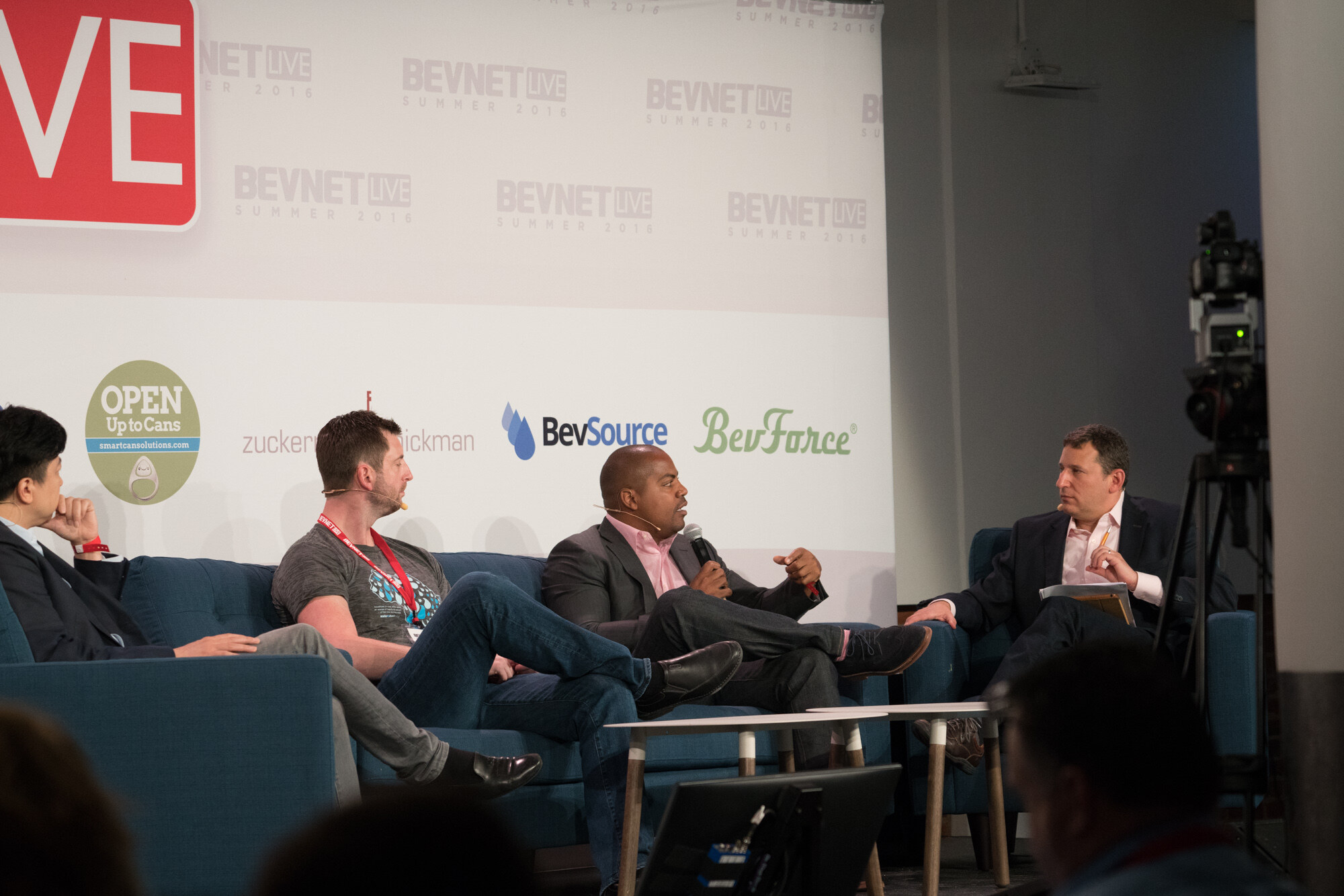 Four men on stage, a moderator in a dark blazer in an armchair at right and three panelists on the sofa, with a video camera on a tripod at right and BevSource and BevForce logos behind.