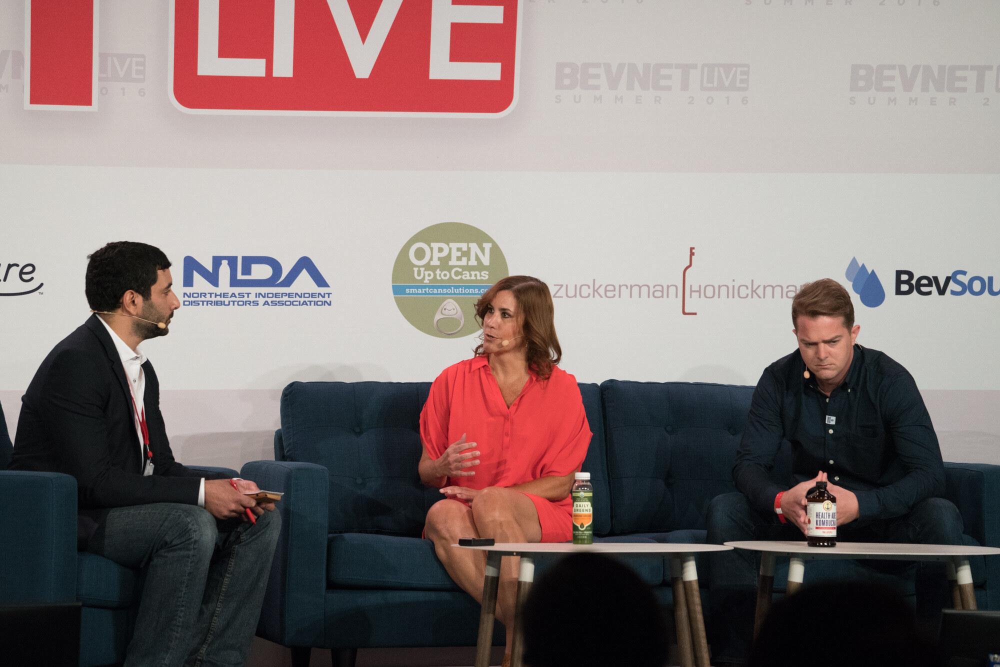 Three people on stage: a moderator in a dark blazer at left, a woman in a coral dress in the middle and a man in a navy shirt holding a Health-Ade Kombucha bottle at right.