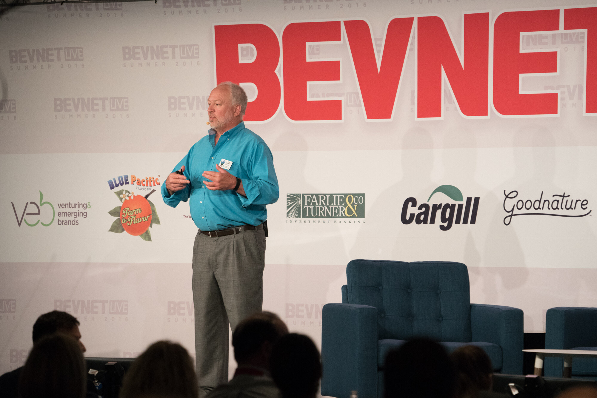 Man in a teal button-up shirt and grey trousers standing on stage, empty armchairs behind him, in front of the red BevNET sign and logos for Veb, Farlie & Turner, Cargill and Goodnature.