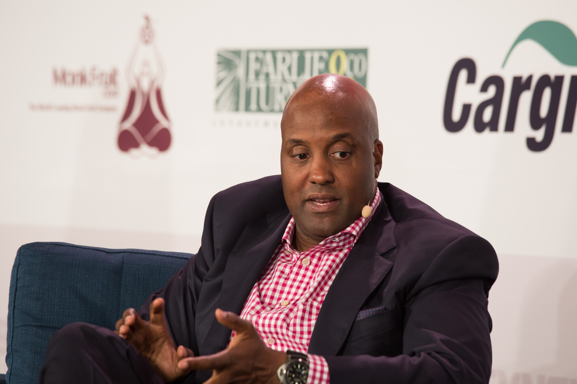 Close view of a man in a dark blazer over a pink-and-white gingham shirt with a headset mic, in front of Monk Fruit Corp, Farlie & Turner and Cargill logos.