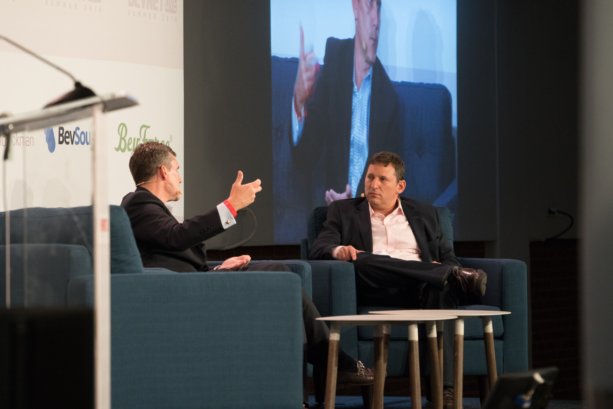 Two men in dark blazers on stage armchairs with a low white table between them, a large screen behind them showing a close-up of a speaker, and a clear podium at left.