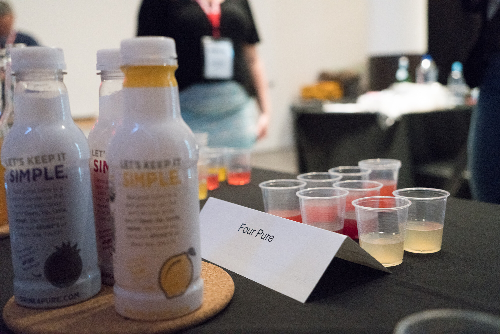 Bottles labelled Let's Keep It Simple on a cork coaster beside a card reading Four Pure and a row of clear plastic tasting cups on a black tablecloth.