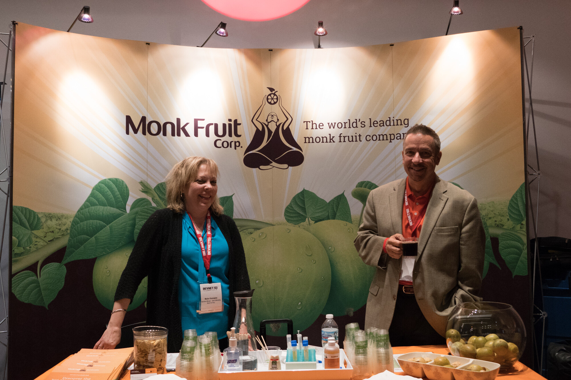 A woman in a teal top and black cardigan and a man in a tan blazer behind an orange table of cups and monk fruit, in front of a Monk Fruit Corp banner.