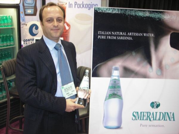 A man in a pinstripe suit and blue tie holds a bottle of Smeraldina water beside a large Smeraldina poster reading Italian natural artesian water, pure from Sardinia.