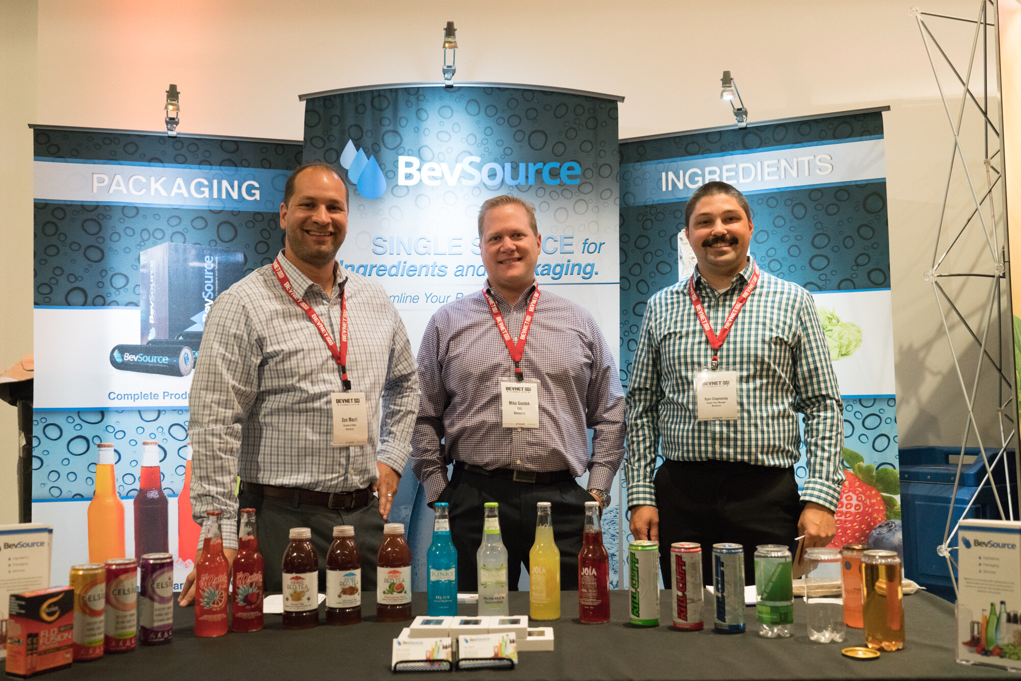 Three men with BevNET Live lanyards behind a black table of bottled drinks and cans, in front of BevSource banners reading Packaging and Ingredients.