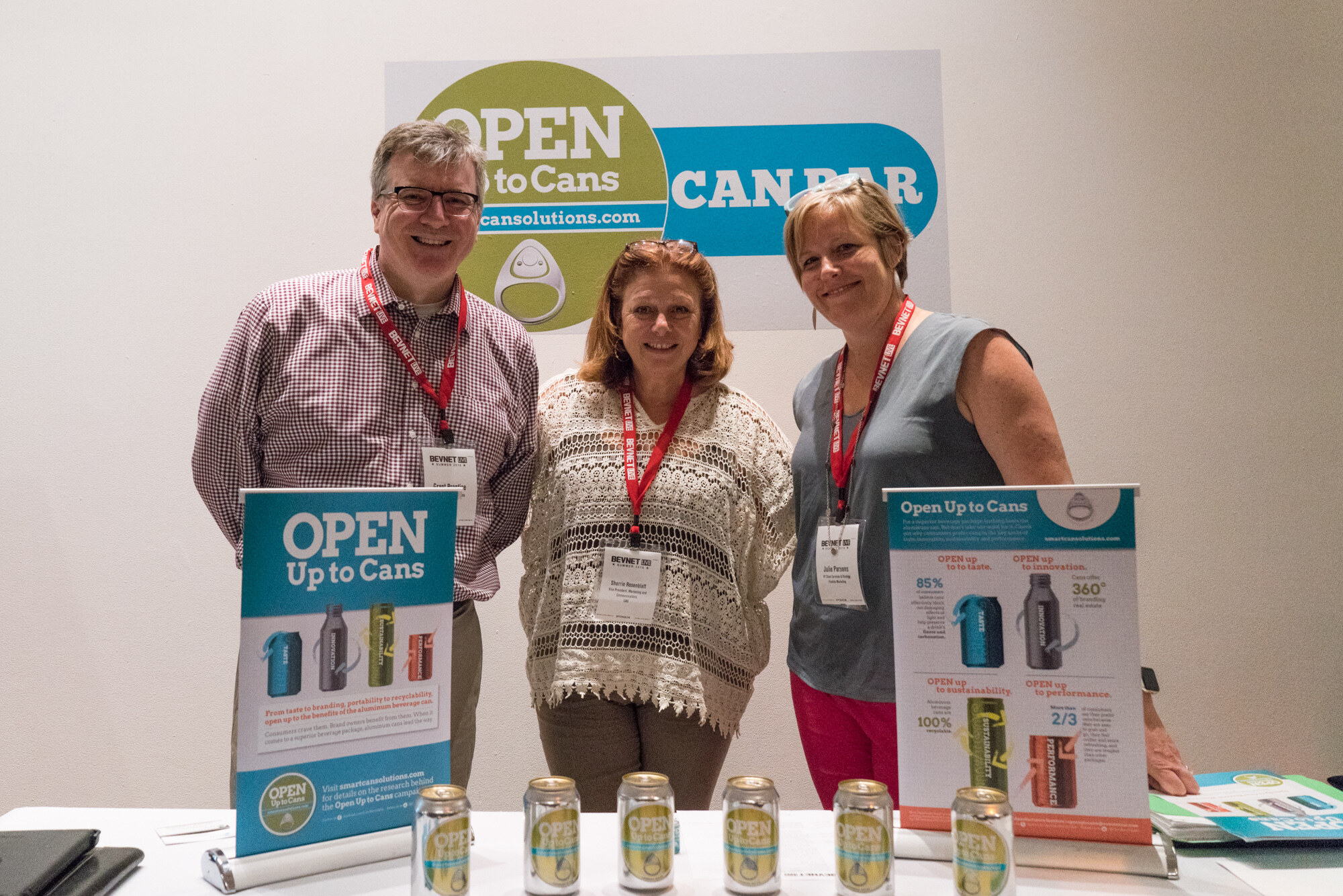 A man in glasses and a checked shirt and two women behind a table with Open Up to Cans banners and cans, under a Can Bar sign.