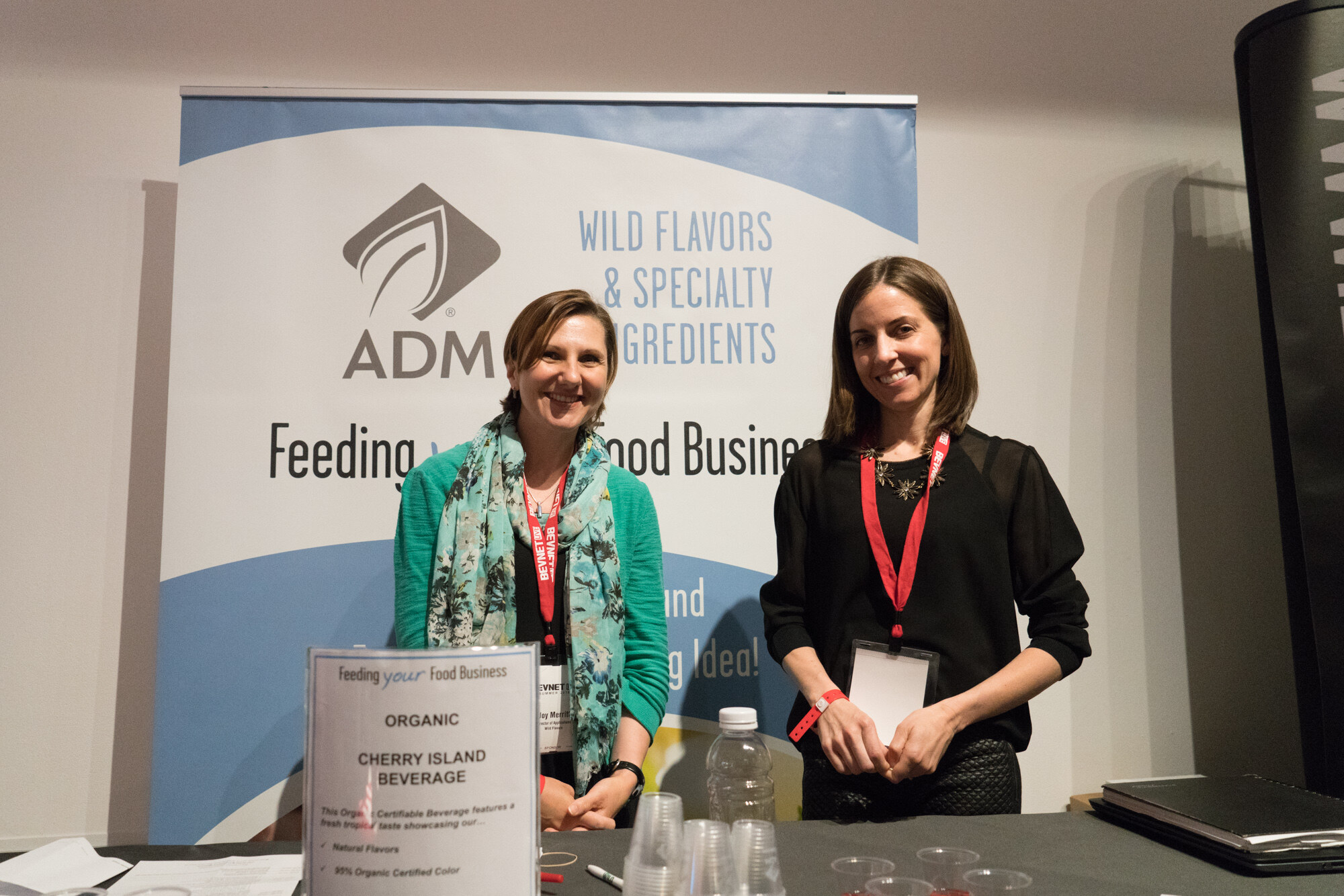 Two women with BevNET lanyards behind a table with a sign for organic cherry island beverage, in front of an ADM Wild Flavors & Specialty Ingredients banner.