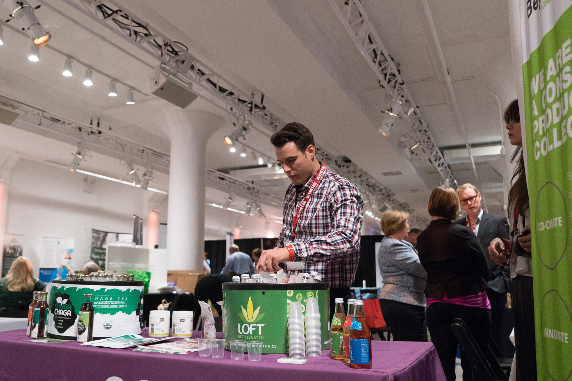 Man in a plaid shirt at a purple-draped table with a LOFT cooler and a Chuga Chaga tea cooler, with attendees and exposed trusses in the background.