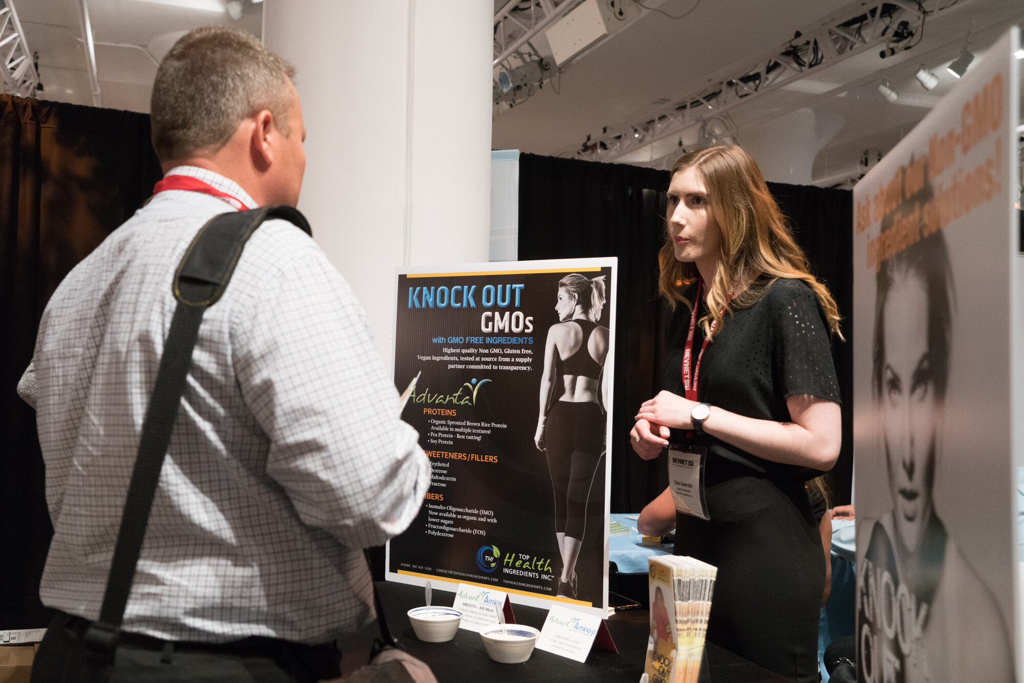 Woman in a black top with a lanyard speaking to a man with a shoulder bag, beside a sign reading Knock Out GMOs with GMO free ingredients, from Advanta.