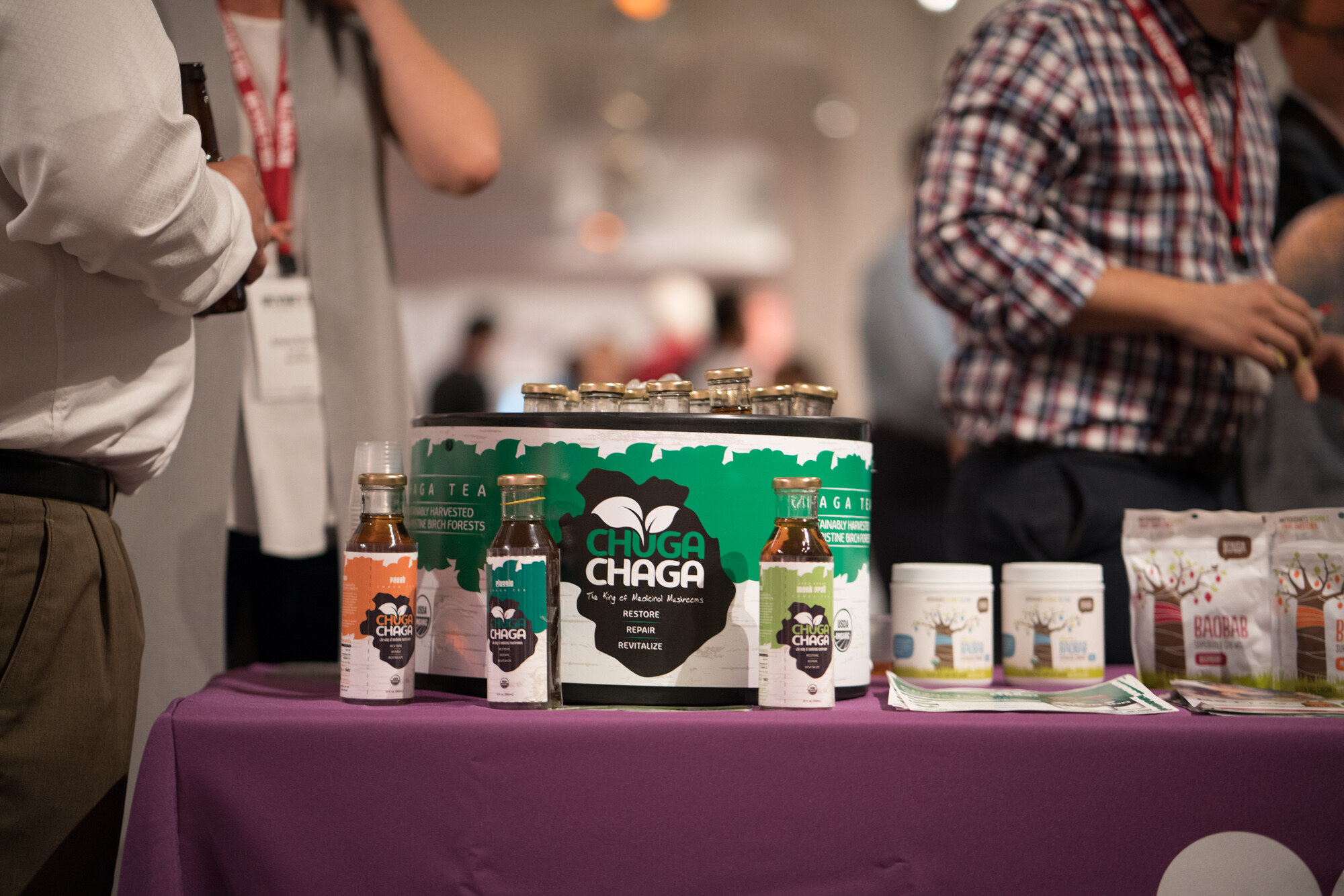 Three Chuga Chaga bottles in front of a green and white cooler, baobab pouches and tubs at right, with attendees' lanyards visible in the blurred background.