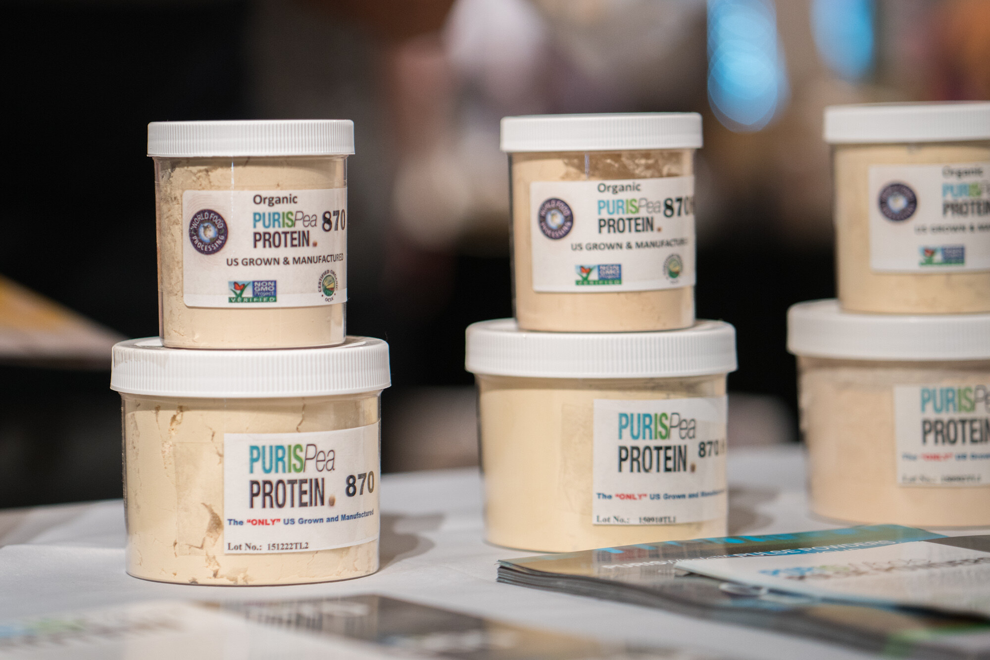 Close view of jars of Organic PURISPea Protein 870 with white lids stacked on a table, with brochures in the foreground and a dark blurred background.