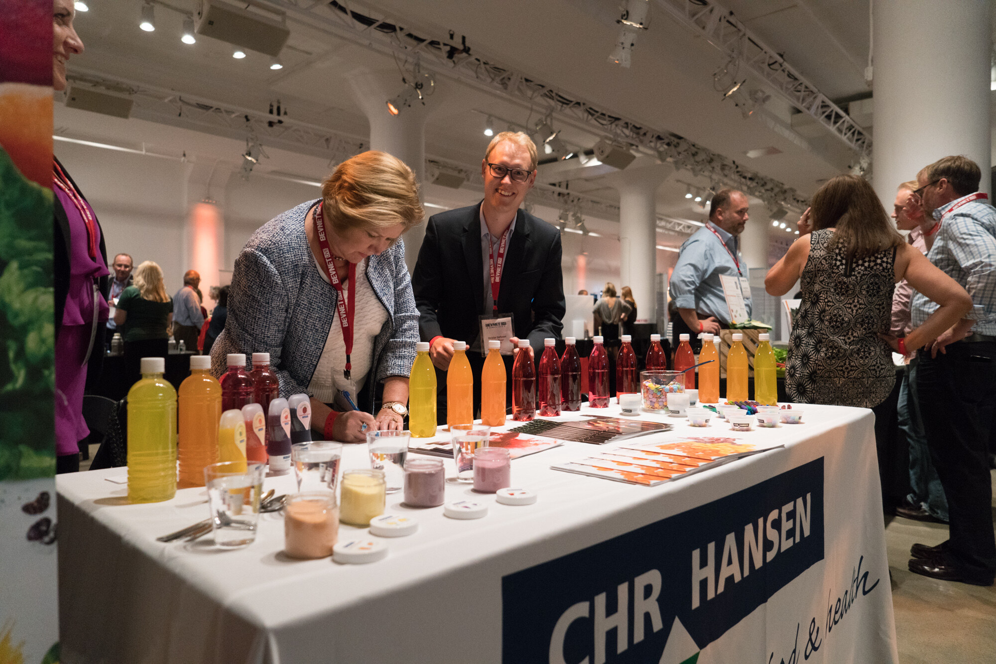 A woman in a tweed jacket writing at a table lined with yellow, orange and red bottles, a smiling man in glasses beside her, and a CHR Hansen table cover.