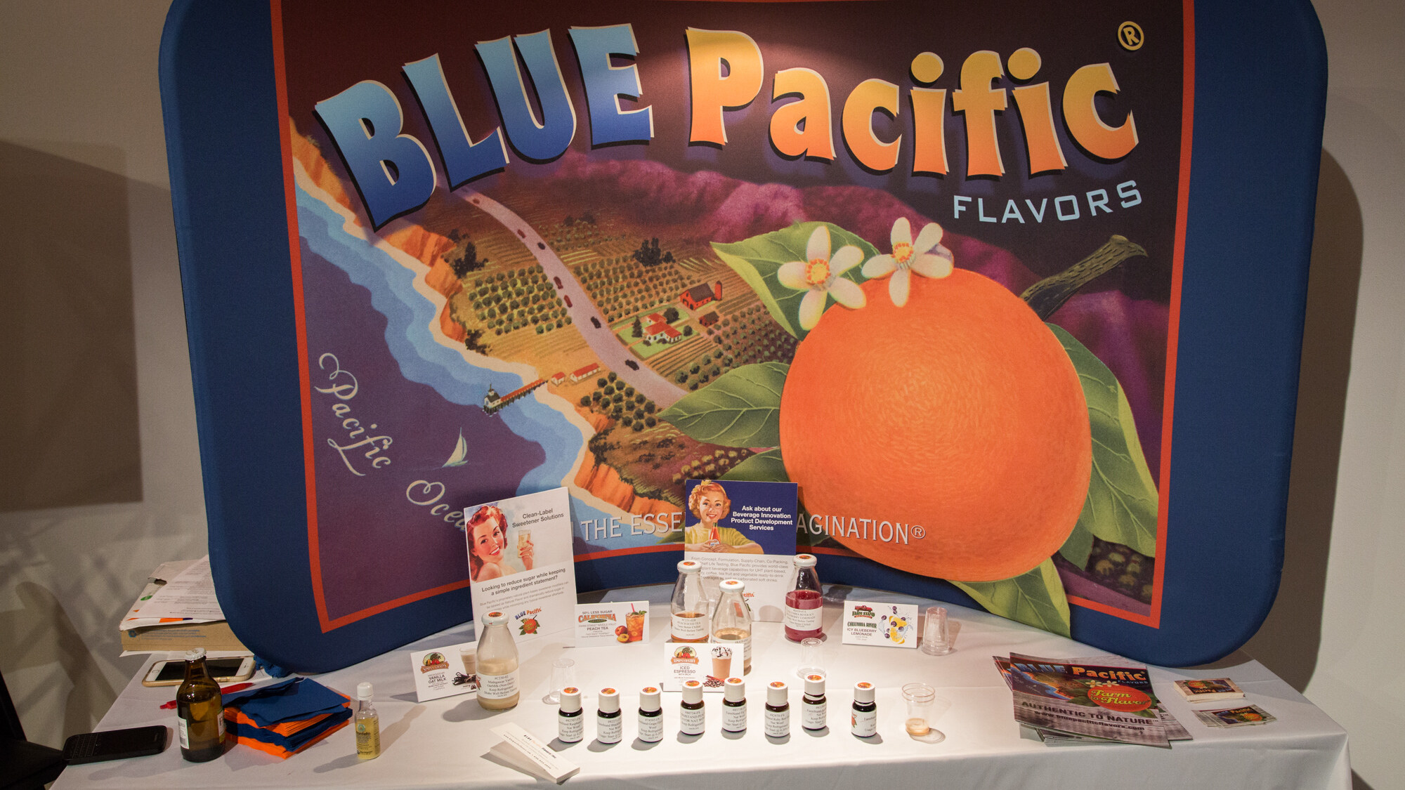 White-draped table with a large Blue Pacific Flavors banner showing an orange and a coastal landscape, small flavor vials, bottles and brochures on the table.