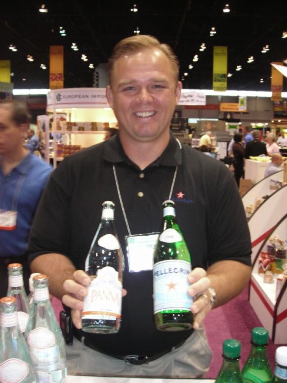 A smiling man in a black polo holds a bottle of Panna in one hand and a bottle of S.Pellegrino in the other. More bottles are on the counter, and trade show aisles are visible behind him.
