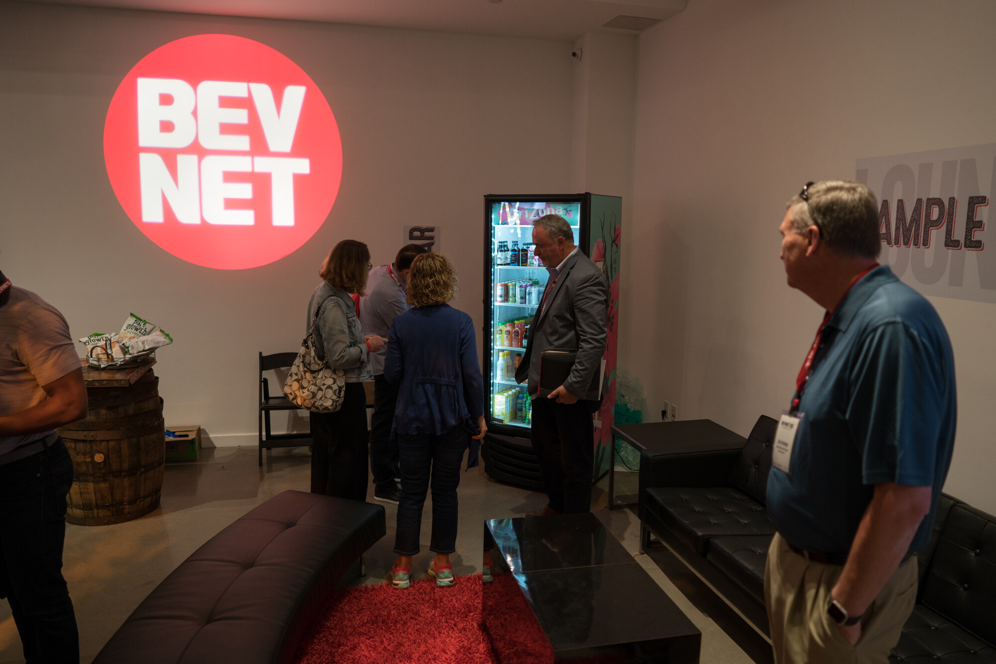 Lounge with a red projected BEV NET logo on the wall, a lit drinks fridge, leather sofas on a red rug and attendees in conversation.