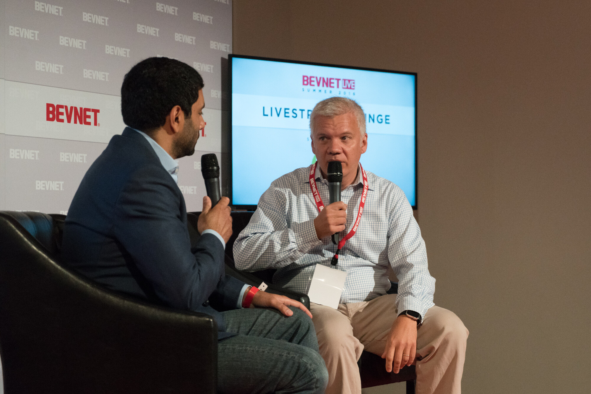 Two men in black leather chairs, one in a navy blazer facing a man in a light checked shirt with a BevNET Live lanyard, with a screen reading Livestream Lounge behind them.
