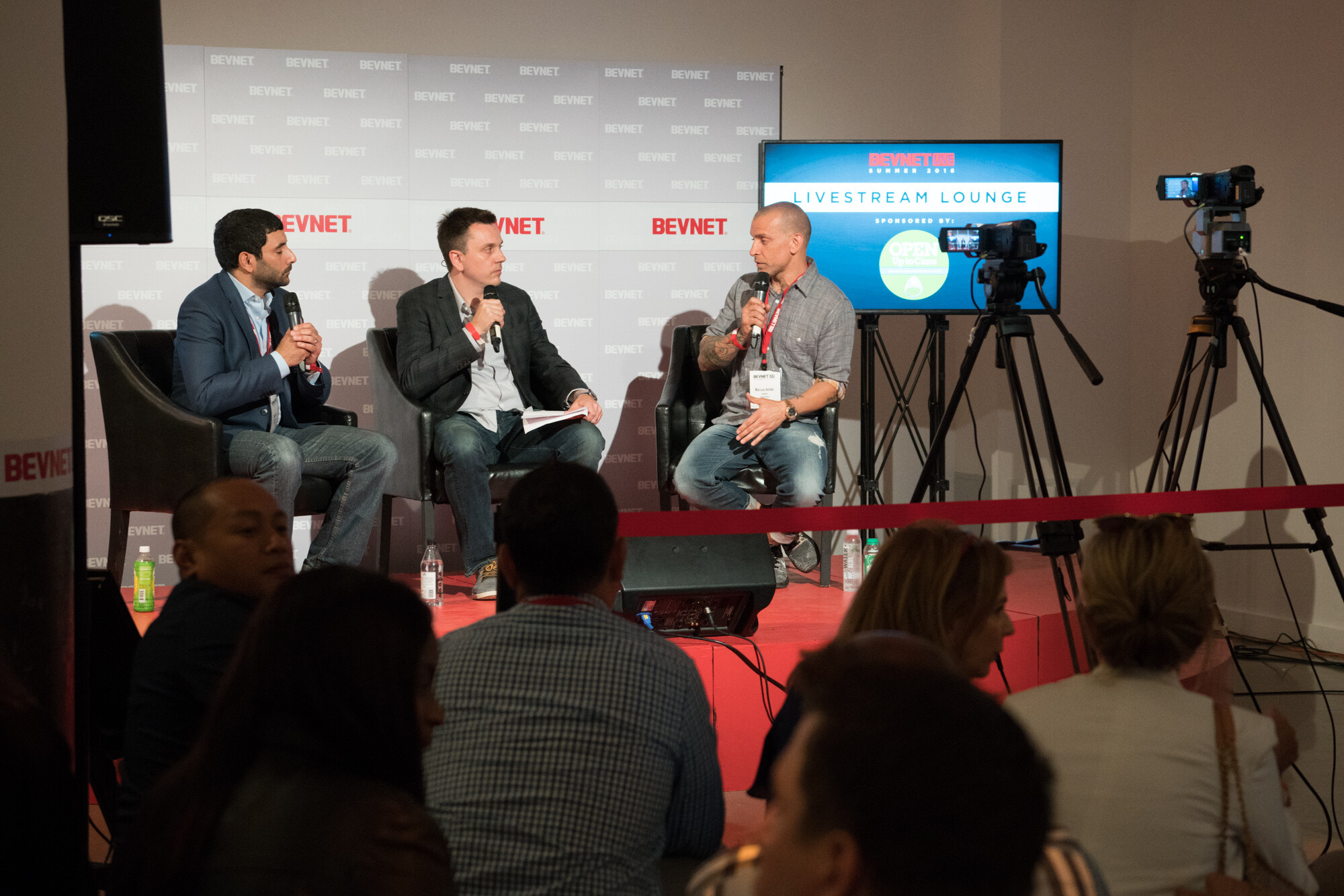 View over the heads of seated attendees toward three people in leather chairs on a red platform, with a Livestream Lounge screen and two video cameras on tripods behind them.