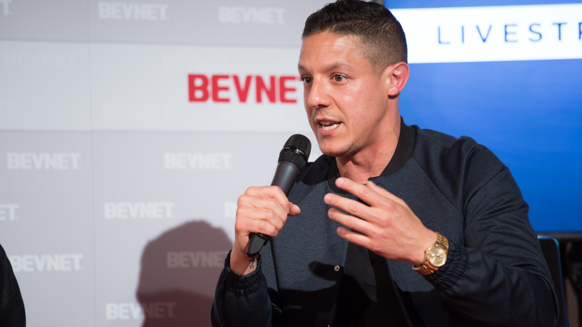 Close view of a man in a navy bomber jacket with a gold watch holding a black microphone, with the BevNET backdrop and the edge of the Livestream Lounge screen behind him.