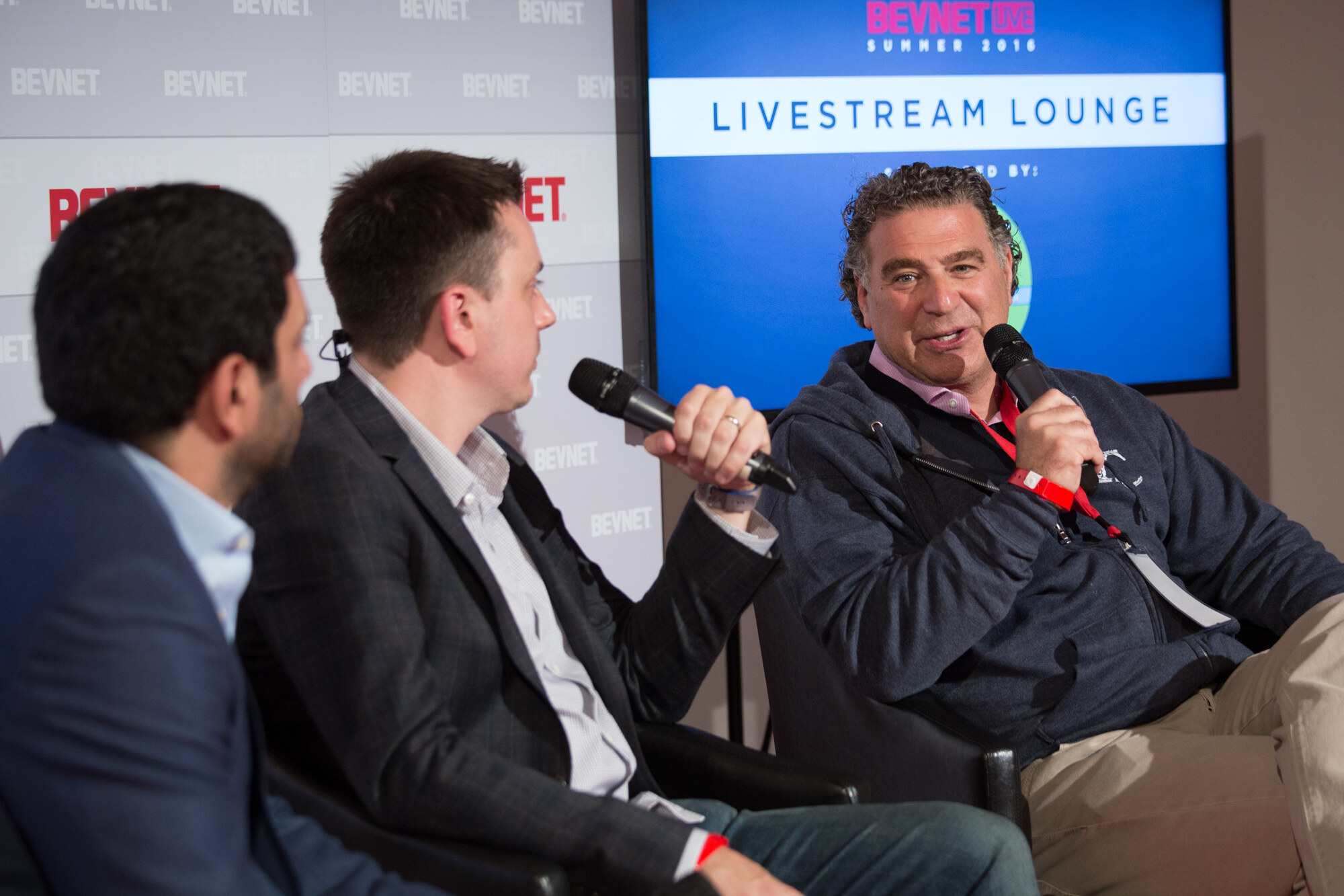 Man with curly hair in a navy quarter-zip speaking into a microphone toward two interviewers, beside a blue screen reading Livestream Lounge, BevNET Live Summer 2016.