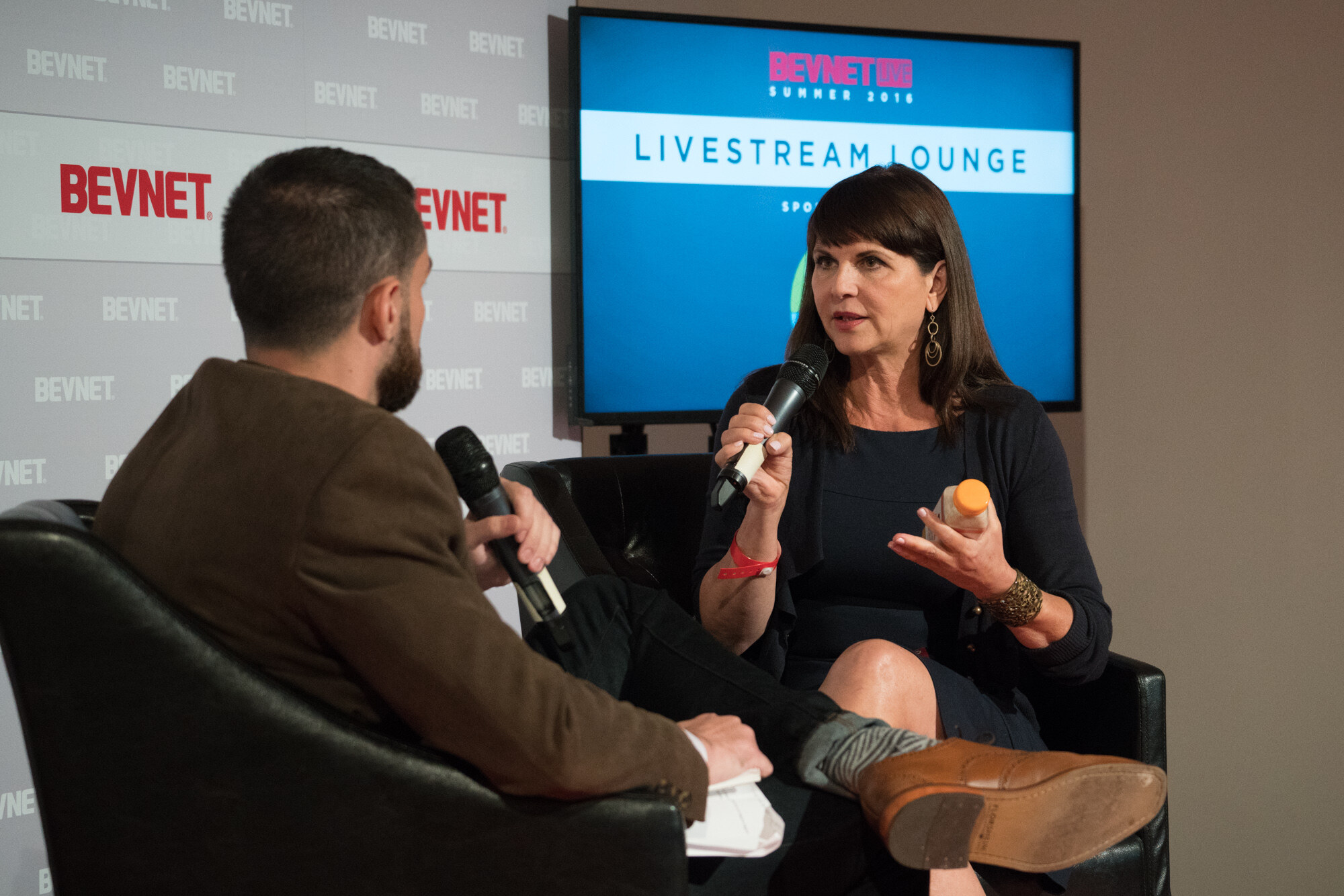 Woman with dark hair in a black cardigan holding a microphone in one hand and an orange-capped bottle in the other, facing an interviewer in a brown jacket, with a Livestream Lounge screen behind.