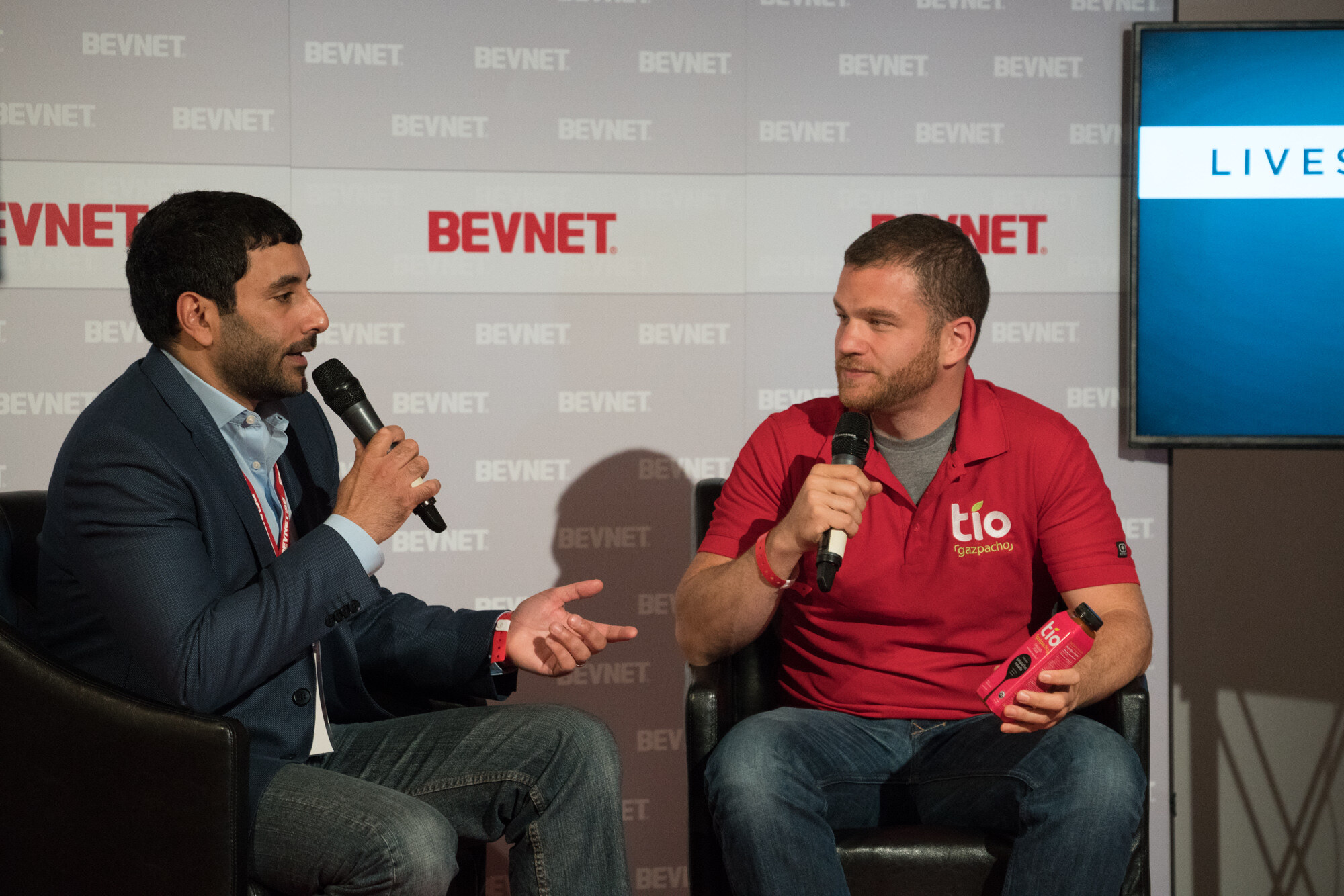 Two men in black leather chairs holding microphones; the guest on the right wears a red polo with a Tio Gazpacho logo and holds a pink bottle with the same logo.
